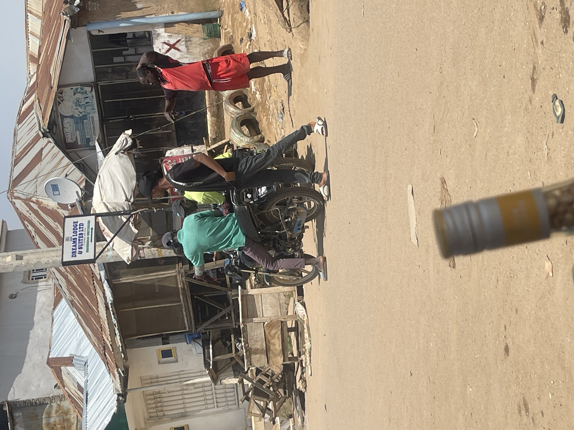 The image depicts a street scene in Abuja, Nigeria. In the foreground, a dirt road or unpaved surface is visible, with some debris scattered on it. A blurred object, possibly a bottle or a camera lens, is in the immediate foreground, obscuring part of the lower section of the image.
Further back, on the right side of the image, a man wearing red shorts and a red shirt stands with his hand on his head, looking to his right. Beside him, also standing, is a man in a black t-shirt and dark pants.
In the middle ground, a motorcycle is parked, and a person wearing a green t-shirt and dark pants is seated on it, facing away from the camera. Another person wearing a bright green shirt is visible behind the motorcycle rider.
The background features several buildings with corrugated metal roofs, some appearing weathered and rusty. One building has a sign that reads "DREAMS LODGE & SUITES LTD" with an arrow pointing to the right. Another building has a sign with "PAULA'S" on it. The overall environment suggests a commercial or residential area in a city. The sky is visible, indicating it is daytime.