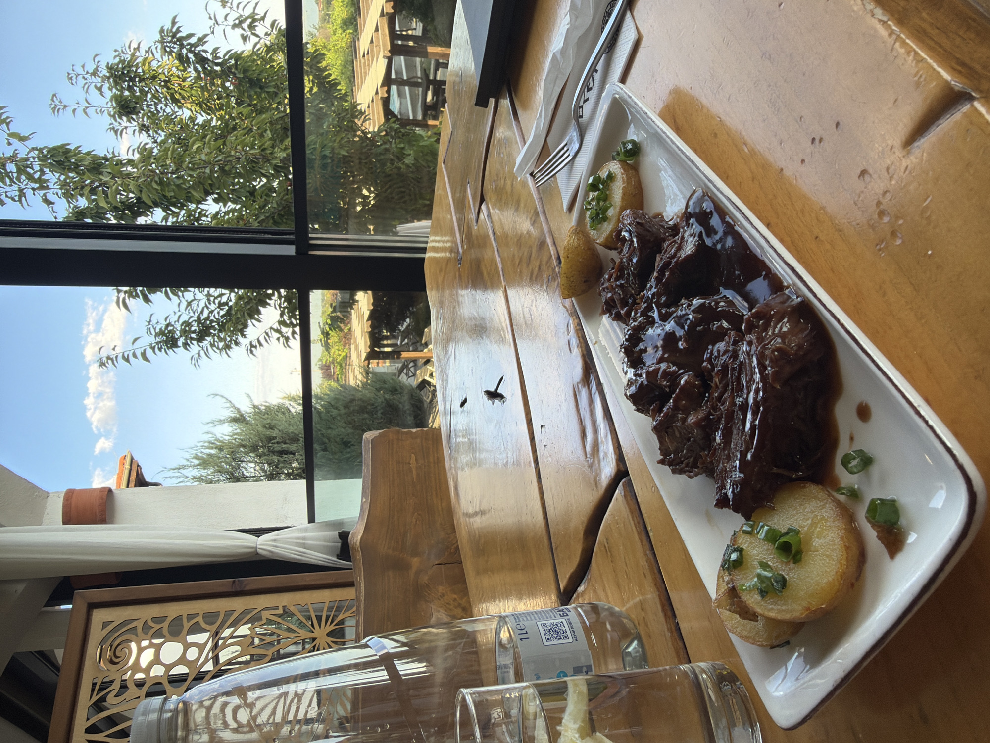 The image shows a meal of braised meat and potatoes served on a white rectangular plate. The plate is positioned on a wooden table, with a glass of water and a bottle of water to the left. In the background, through a large window, a view of an outdoor seating area with pergolas and lush greenery is visible. The scene appears to be from a restaurant in Krasnovo, Bulgaria, given the mention of the location. The lighting suggests it is daytime, likely late afternoon or early evening, with the sun casting warm tones on the wooden surfaces. The overall atmosphere is relaxed and inviting, suggesting a pleasant dining experience.