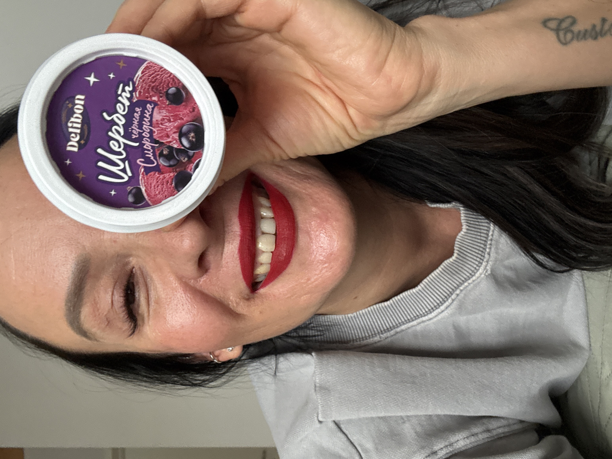 A woman is holding a container of Delibon black currant sorbet in front of her face. The woman has dark hair and is wearing red lipstick. She is wearing a light gray sweatshirt. The brand name on the sorbet container is in Cyrillic. The image was taken in Chaikovsky, Russia.