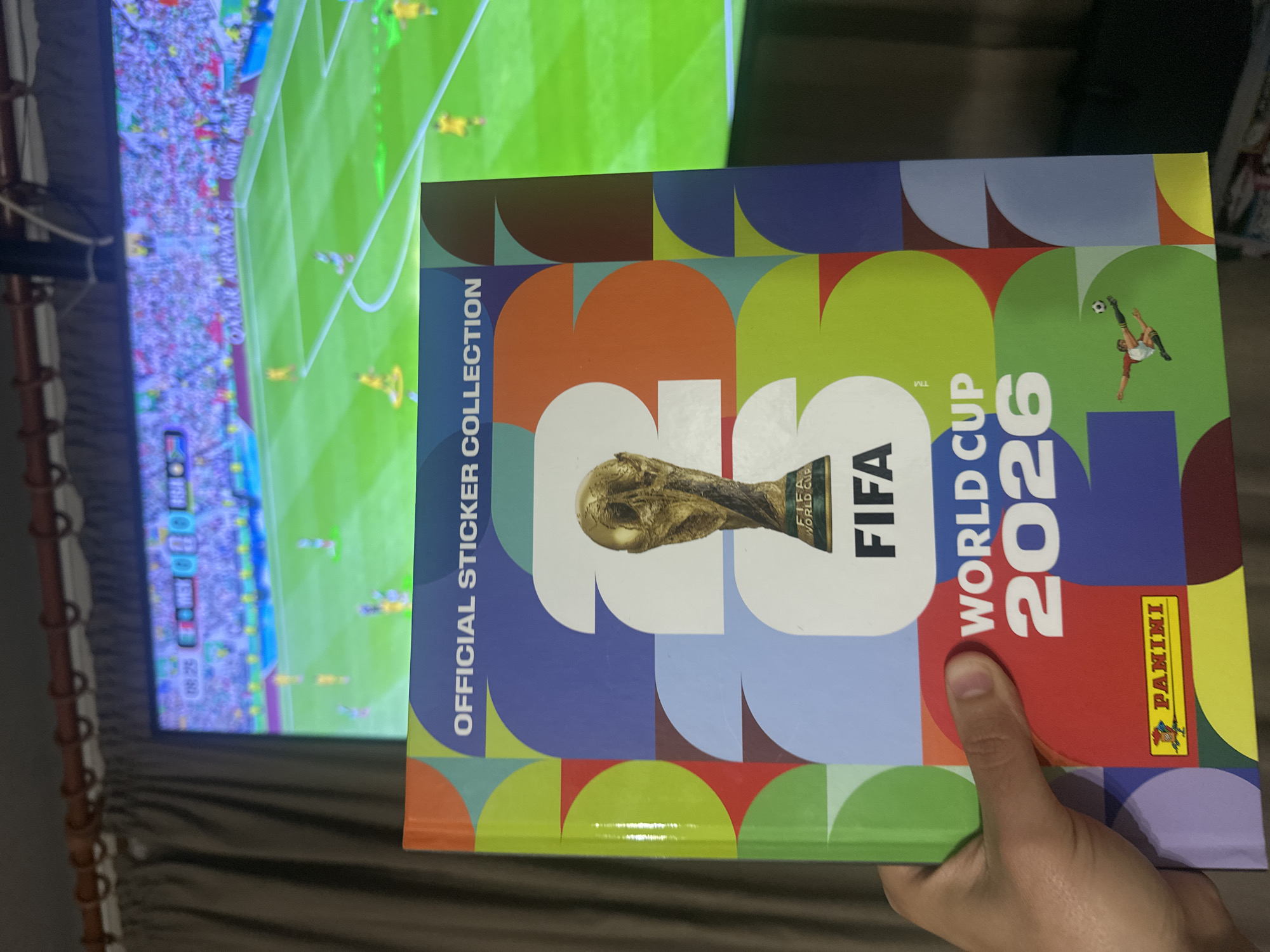 In an indoor setting, a person's hand is holding a "FIFA World Cup 2026 Official Sticker Collection" album. The album features a prominent image of the FIFA World Cup trophy and the text "FIFA WORLD CUP 2026." A television screen in the background displays a soccer match with visible game statistics. The environment suggests a home entertainment area with curtains and what appears to be a cable or internet box mounted above the television. The scene does not contain any elements that indicate the city of Prisovo, Bulgaria.