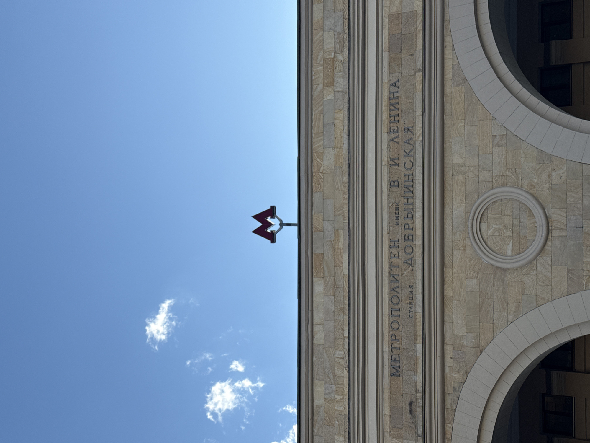 This is a low-angle shot of a metro station entrance in Moscow, Russia, under a clear blue sky with a few wispy clouds. The image is oriented vertically, with the sky on the left and the building facade on the right.
The building is made of light-colored stone blocks and features fluted columns and arched openings. A prominent red metro symbol is visible against the sky. On the facade, there is inscribed text in Russian. The text reads "МЕТРОПОЛИТЕН ИМЕНИ В. И. ЛЕНИНА" which translates to "Metro named after V. I. Lenin". Below this, it says "СТАНЦИЯ ДОБРЫНИНСКАЯ", meaning "Dobryninskaya Station". The style of architecture suggests a Soviet-era building. The lighting indicates it is daytime, likely a sunny day. There are no people visible in the frame. The overall impression is one of civic architecture and public transportation infrastructure.