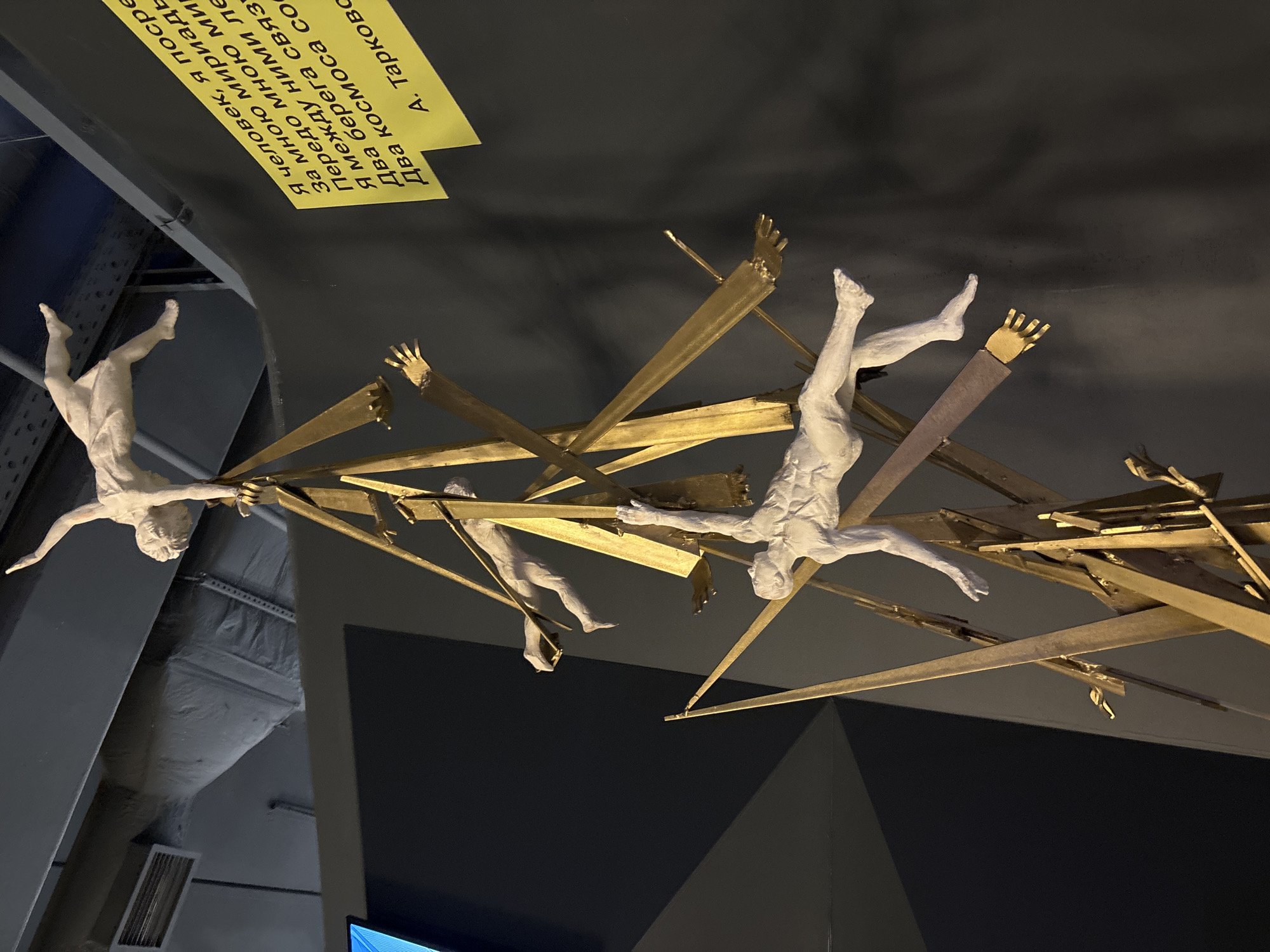 The image depicts a sculpture in Saint Petersburg, Russia. The sculpture features multiple human figures in white, seemingly suspended or falling, interspersed with golden metallic beams or supports. A plaque with text in Russian, attributed to A. Tarkovsky, is visible near the top of the frame. The background consists of gray walls, with architectural elements such as beams and a ventilation system subtly visible. The environment suggests an indoor exhibition space.