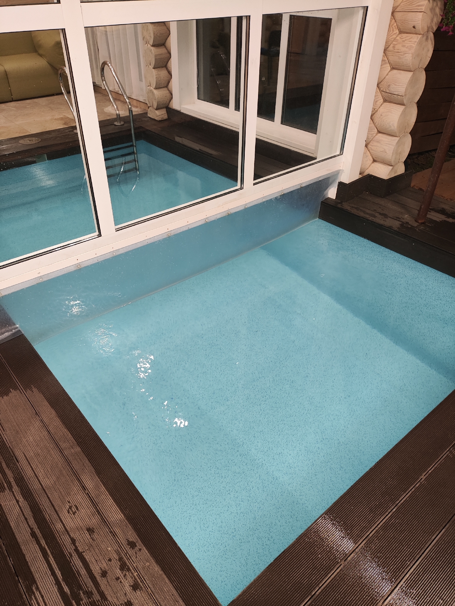 This media file captures an indoor swimming pool area with a rustic, cabin-like aesthetic. The pool itself is filled with clear, light blue water, appearing inviting and serene. The bottom of the pool has a speckled texture. A stainless steel ladder with a curved handle is partially submerged, indicating the pool is ready for use.
The pool is surrounded by a wooden deck made of dark brown planks, some of which appear wet, suggesting recent use or the humid environment of a pool area. To the left of the pool, large glass windows with white frames offer a view into another part of the building, where a light-colored sofa and a tiled floor are visible.
The structure of the building is notable, with a prominent wall constructed from rounded logs, characteristic of a log cabin. This log wall, with its hexagonal patterns where the logs meet, is a strong visual cue to the location's style. White window frames and metal accents are visible.
The lighting suggests it could be evening or an overcast day, as there are no strong shadows indicating direct sunlight. The overall atmosphere is peaceful and relaxing, hinting at a private retreat or a spa-like setting. There is no visible text in the image. The context provided, Старое Мартьяново, Russia, reinforces the impression of a cozy, potentially rural or resort-like environment.