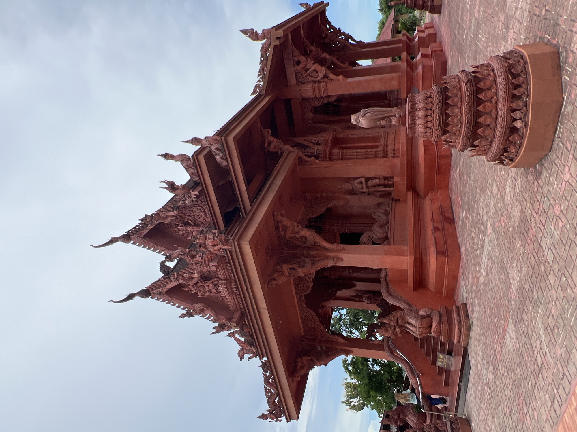 The image shows the exterior of a red temple structure in Baan Hua Thanon, Thailand. The temple features intricate carvings and ornate roof designs, typical of Thai architecture. The building is constructed from red-colored material, likely concrete or stucco, and is adorned with numerous sculptures of mythical creatures and figures. In the foreground, a tiered, conical red sculpture sits on a patterned red brick walkway. To the left of the temple, a staircase with dragon-like railings leads up to the entrance. A person with blonde hair is visible walking up the stairs. Trees and other structures can be seen in the background under a cloudy sky. The overall impression is one of a visually striking and culturally significant religious site.