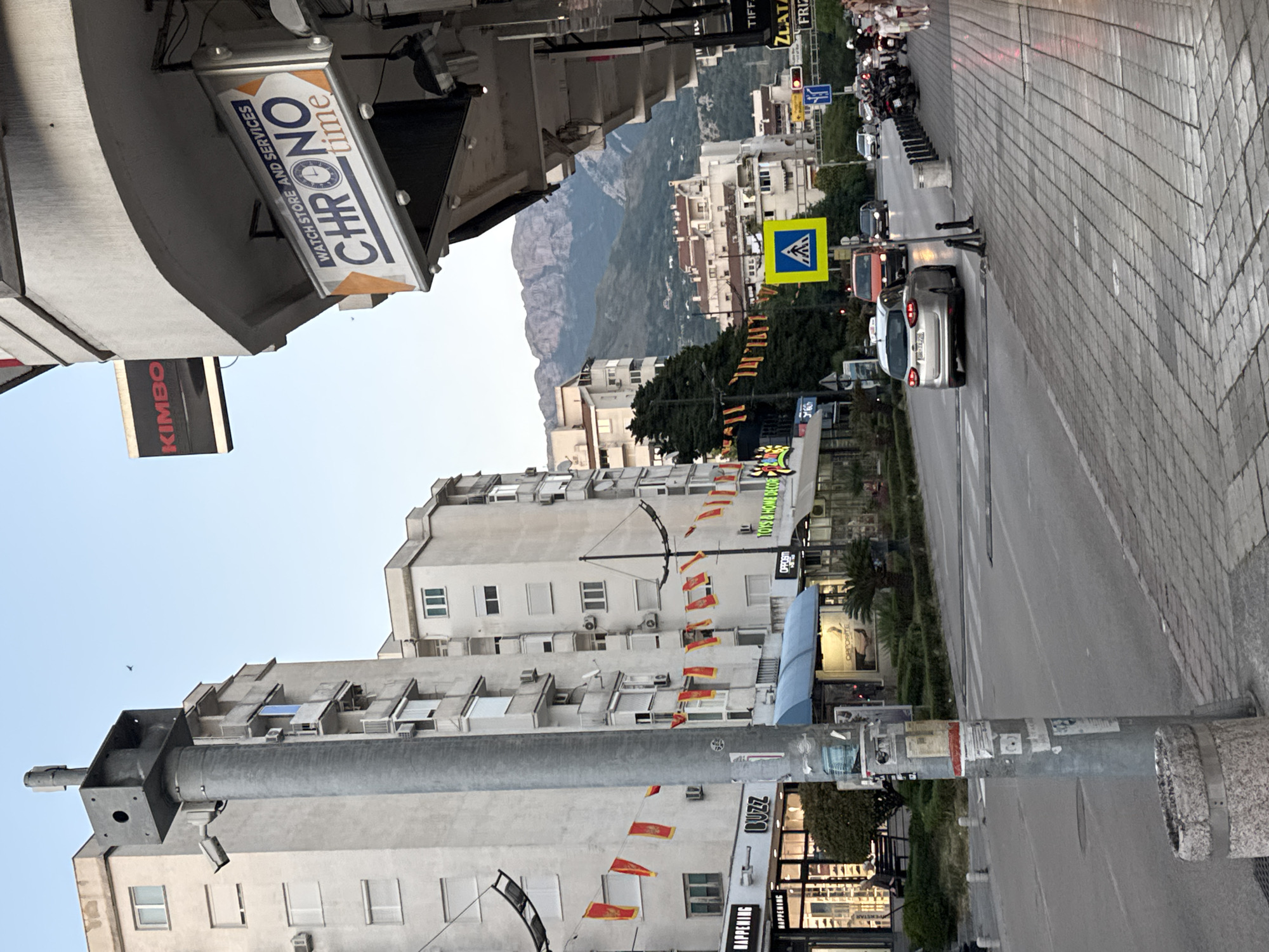 The image shows a street view in Bar, Montenegro. Buildings line the street, adorned with flags. Signage for various businesses like "BUZZ," "TOYS & HOME DECOR," "OPPOSM," "HAPPENING," "KIMBO," and "CHRONO time WATCH STORE AND SERVICES" are visible. There is a street pole in the foreground, plastered with stickers and notices. A crosswalk with a pedestrian sign marks the road, and cars and motorcycles are present in the traffic. In the background, a mountain range is visible under a clear sky.
