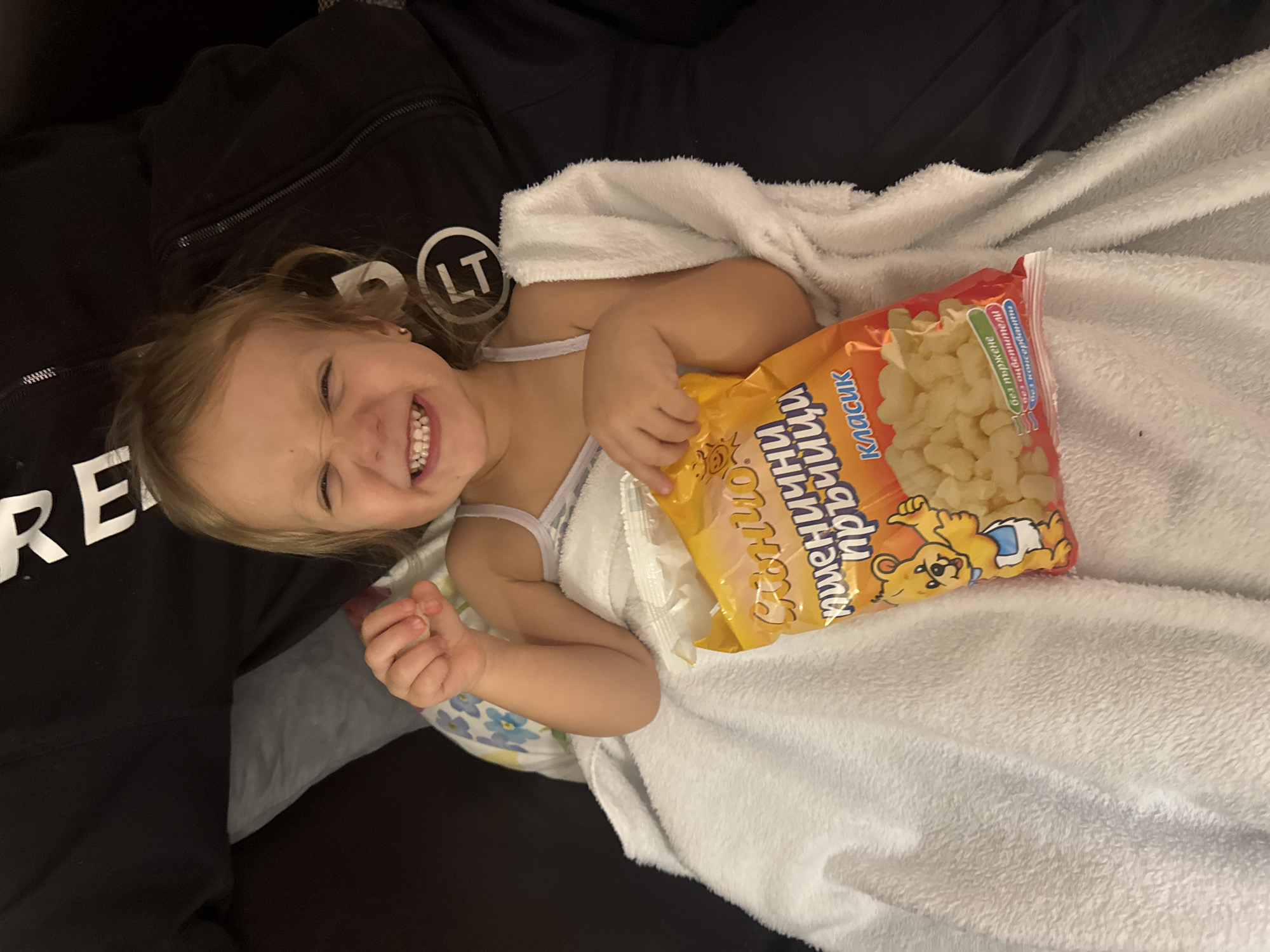 A young child with fair hair is lying on their side, wrapped in a white blanket, and smiling broadly. They are holding a yellow and red package of "Puffed Prince" snacks, with Cyrillic writing and an image of a bear wearing a diaper, seemingly on a lap or surface covered in black clothing. The context suggests an indoor setting, but the specific location in Varna, Bulgaria, cannot be determined from the image alone.