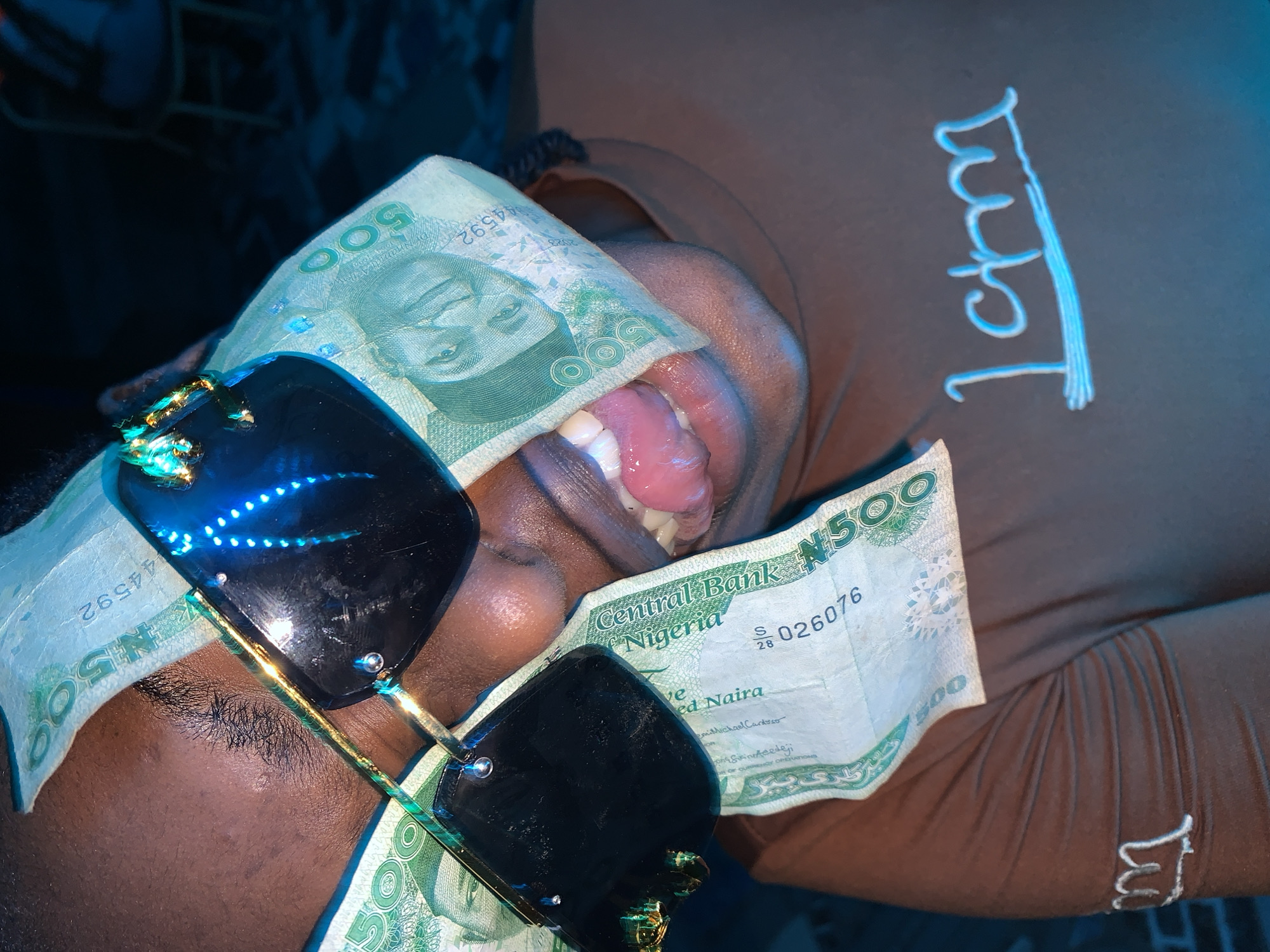 A close-up, high-angle portrait depicts an individual, likely male, with dark skin, posing in an indoor setting within the city of Kabong, Nigeria. The subject's face is partially obscured by dark-lensed sunglasses with gold-toned frames, under which multiple Nigerian 500 Naira banknotes are carefully tucked, covering portions of the forehead and cheeks. The subject's mouth is open, revealing white teeth and a slightly extended pink tongue. The individual wears a brown short-sleeved shirt featuring a light blue, underlined inscription, possibly "1ctm," on the chest/shoulder area. The scene is characterized by a strong blue ambient light, which reflects off the sunglasses and illuminated areas of the skin. The background is dark and blurred, with faint details of a patterned floor and a light-colored chair visible in the distance.