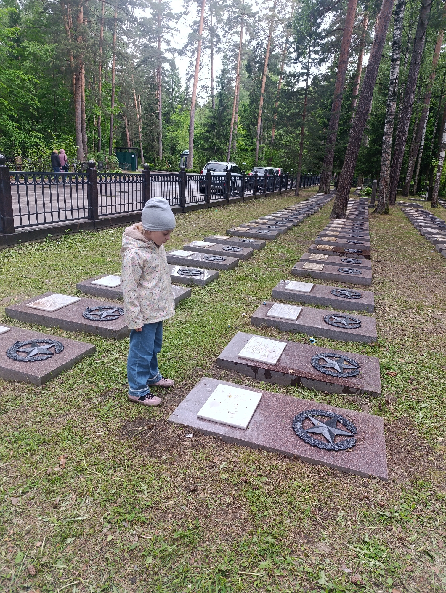 A young child stands in a cemetery in Noginsk, Russia. The cemetery contains rows of identical grave markers with a star emblem. The child is wearing a gray hat, a light-colored jacket, and jeans. Tall trees surround the cemetery. A wrought-iron fence and cars are visible in the background.