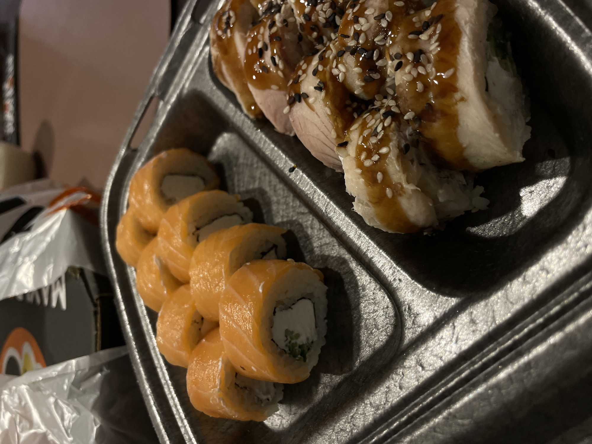 A top-down, close-up shot shows a grey disposable food container divided into two sections. The container is filled with sushi rolls. In the left section, salmon-covered rolls are visible, revealing white cream cheese and small pieces of green filler inside. In the right section, white-meat rolls are drizzled with a brown sauce and sprinkled with white and black sesame seeds. The left part of the image shows a small part of packaging. The photograph does not indicate the location of Sepych, Russia.