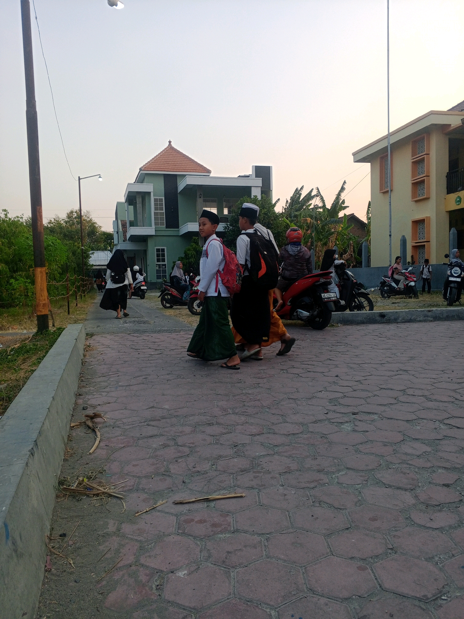 In Lembah, Indonesia, a paved walkway with hexagonal stones leads towards a building complex. On the right, a light yellow building with a brown roof and decorative window openings stands. To the left, a green building with a similar brown roof and modern architectural features is visible. The sky is overcast and light. In the foreground, a concrete curb lines the left side of the walkway. Two boys, dressed in traditional Islamic attire with white shirts, black caps, and long green skirts, are walking towards the right. One of them carries a red backpack. Other individuals and motorbikes are present in the mid-ground. Trees and foliage are visible behind the buildings, suggesting an outdoor setting, possibly near a school or religious institution.