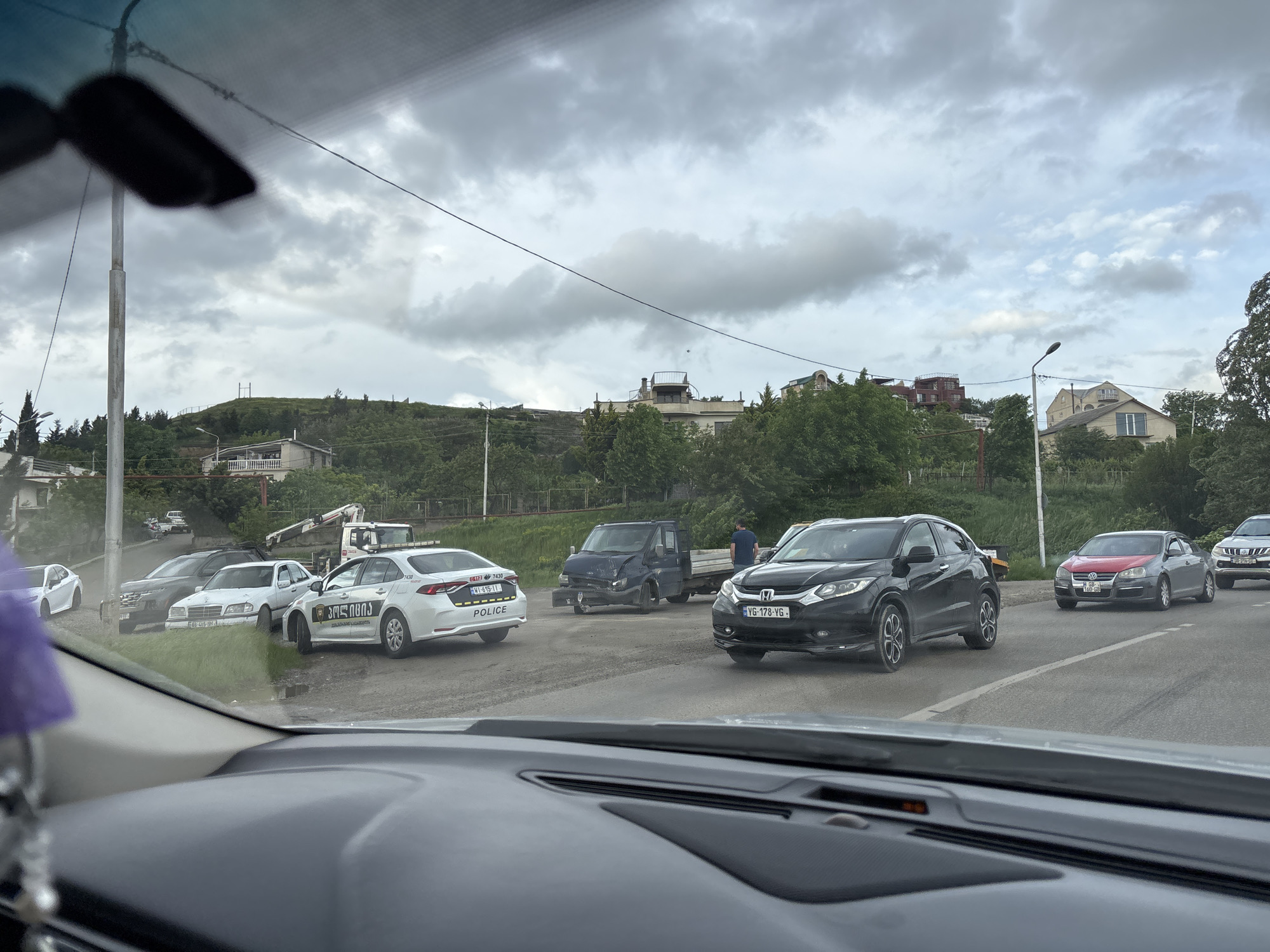 The image depicts a street in Tbilisi, Georgia with several vehicles, including a black Honda HR-V with license plate "VG-178-YG," a white police car with Georgian script and license plate "WI-615-II," a damaged blue flatbed truck, a grey Volkswagen Jetta with a red hood, and several parked cars along the side of the road. A person is standing near the flatbed truck. The image is taken from inside a vehicle, looking out through the windshield. The sky is overcast. Buildings and vegetation are visible on a hill in the background.
