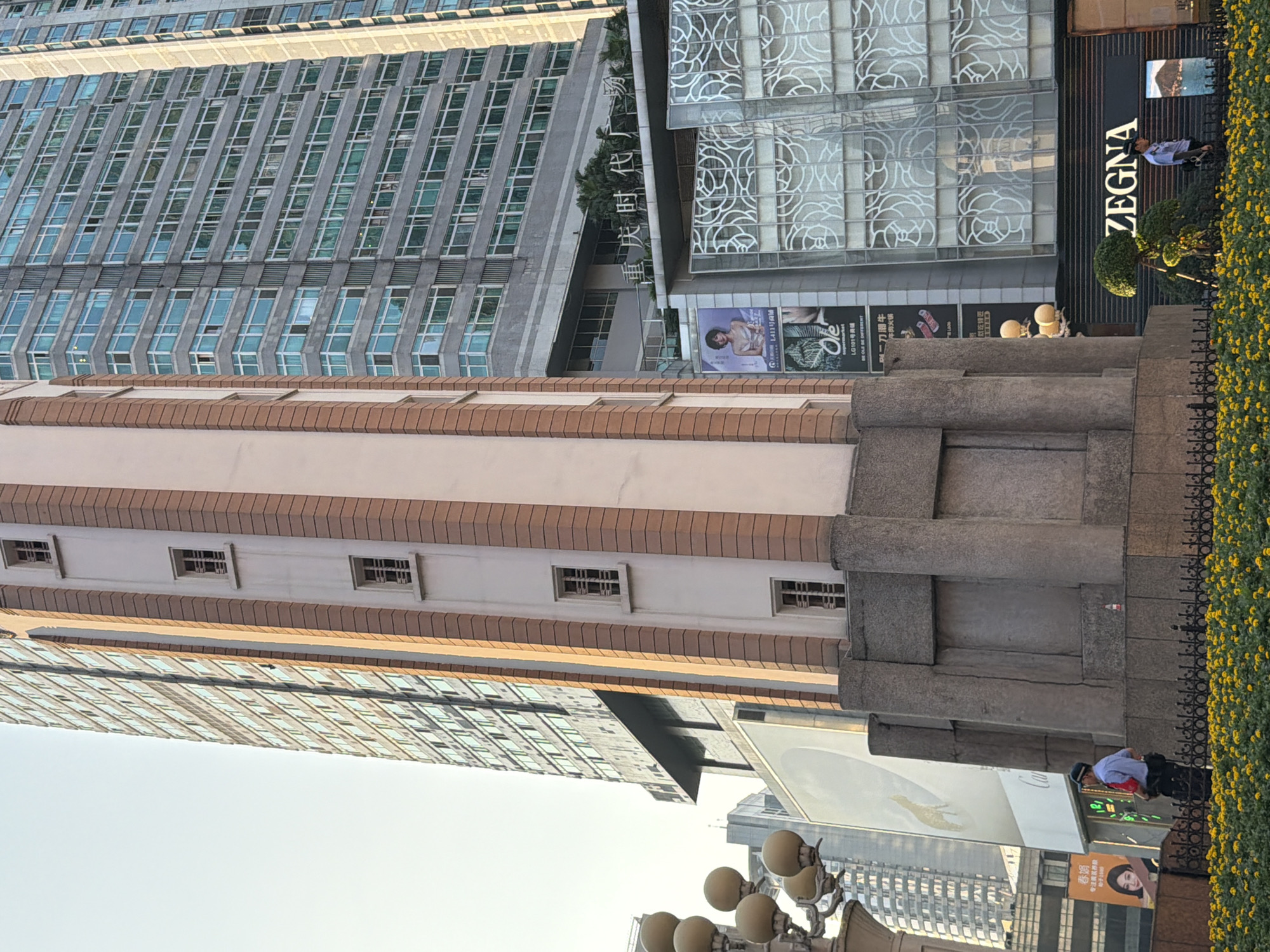 The image captures a view of a cityscape in Yuzhong District, China, focusing on a tall, modern building with a grey facade and numerous windows. Adjacent to it, a narrower, lighter-colored structure with vertical brown accents and smaller, framed windows is visible. In the foreground, a person dressed in a light blue uniform and a dark cap, possibly a security guard, stands facing right. They are wearing a dark belt with a pouch and have a red item tucked into their waistband. Behind the guard, a black metal fence encloses a bed of vibrant yellow flowers. The guard is positioned in front of a concrete structure with large, recessed panels. Above the concrete structure and to the right, a building features a prominent sign with the word "ZEGNA" in large white letters on a dark background, with a decorative green topiary in front of it. Further to the right, a man in a similar uniform is walking. A large billboard with Chinese characters and an image of a woman in a dress is displayed on a lower section of the building to the left. The sky is clear and bright.