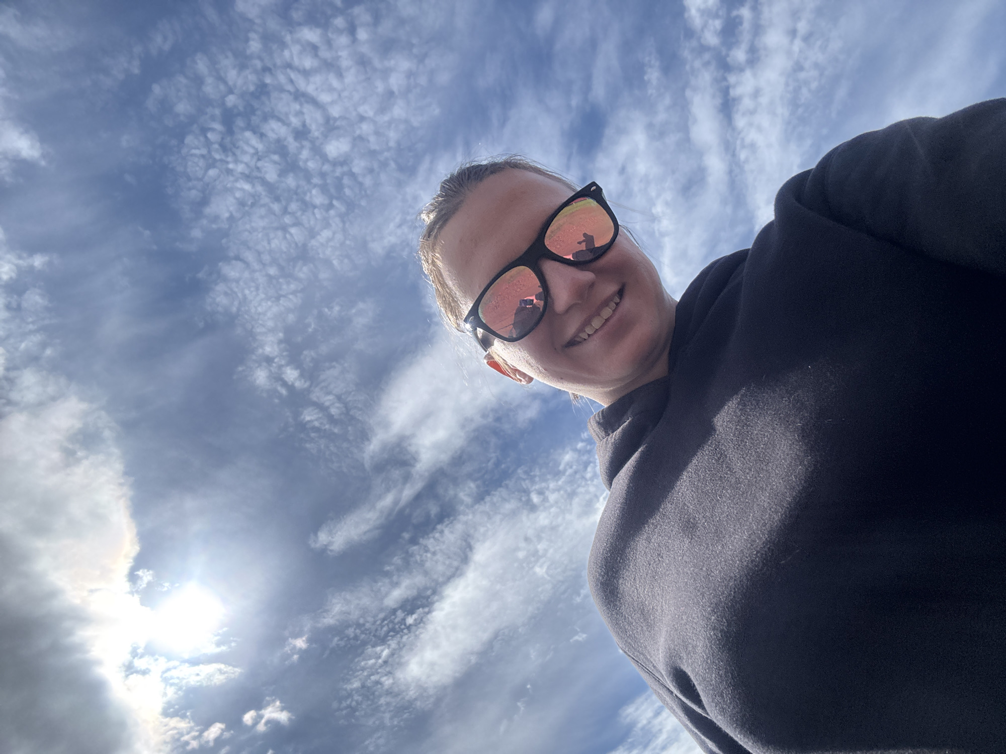 A low-angle selfie captures a woman with fair skin and light hair pulled back, wearing black sunglasses with orange reflections, and a dark hooded sweatshirt. She is smiling. The background is a bright, blue sky filled with altocumulus clouds and the sun is visible through a gap in the clouds. It is not possible to determine the location is in Раздолье, Russia from the image.