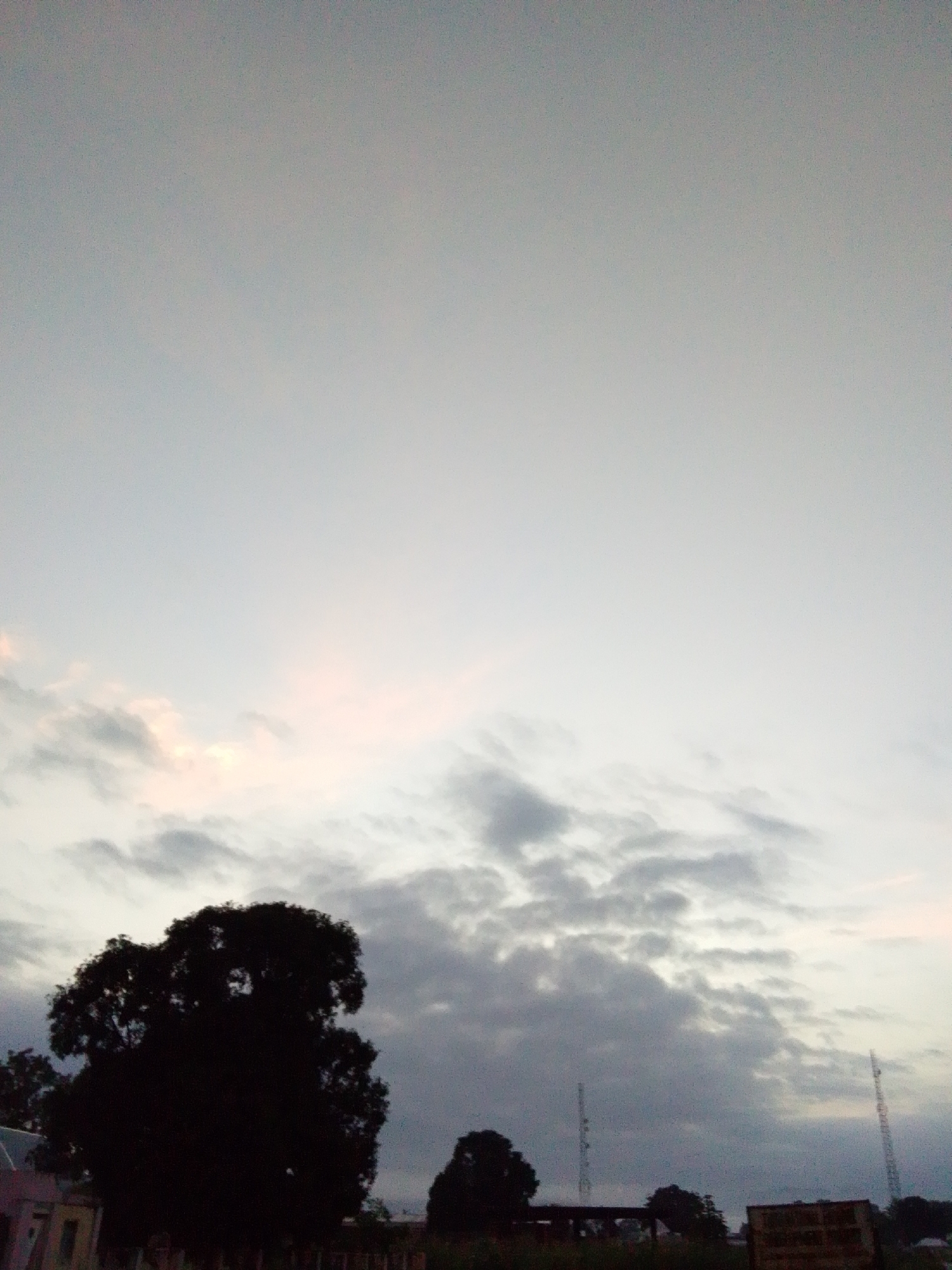The image depicts a twilight scene in Serti, Nigeria, dominated by a vast, overcast sky with scattered clouds. The clouds are a mix of dark grays and lighter, possibly pinkish hues, suggesting the remnants of sunset or the approach of dawn. In the foreground, a large, silhouetted tree with a dense canopy takes up the left portion of the frame. To its right and further in the distance, two slender communication towers pierce the sky. The ground level reveals the faint outlines of buildings and possibly other vegetation, obscured by the low light and shadows. The overall atmosphere is serene and somber, characteristic of the transition between day and night.