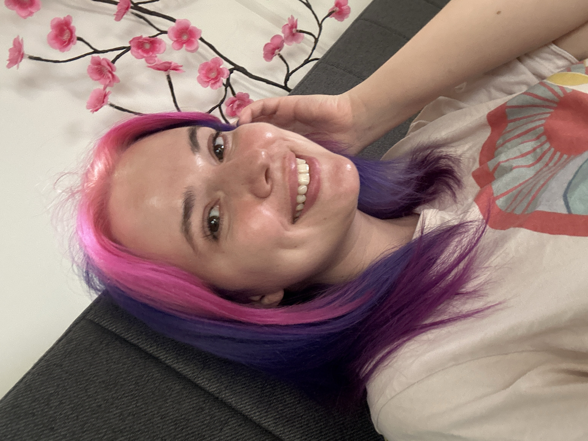 A woman with pink and purple hair is lying on a gray couch. There are pink cherry blossoms on the wall behind her. She is smiling. She is wearing a light-colored t-shirt. The location cannot be determined from this image.