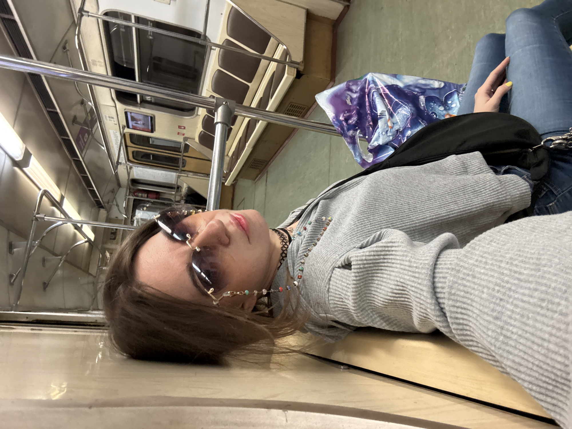 A woman wearing sunglasses, a gray sweater, and blue jeans is lying down in a Moscow Metro train car. Her head rests on a light-colored surface, possibly the top of the train's interior wall. A black fanny pack and a purple shopping bag are next to her. Brown leather seats are visible in the background.