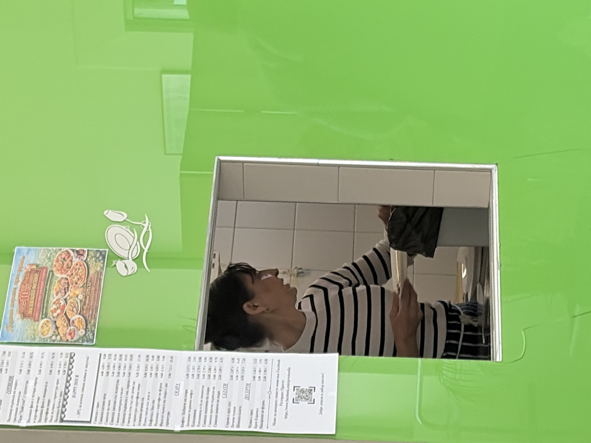 This is an interior shot of a food establishment, likely a restaurant or cafe, in Sofia, Bulgaria, as suggested by the Cyrillic text on the menu. The scene is viewed through a serving hatch or window, characterized by a bright green surround. The menu is affixed to the green surface, displaying a range of food items and prices in Bulgarian Lev (лв). Adjacent to the menu is a decorative sticker featuring a culinary-themed design. Through the serving hatch, a person with dark hair is visible, wearing a striped top. The interior beyond the hatch is characterized by white-tiled walls.