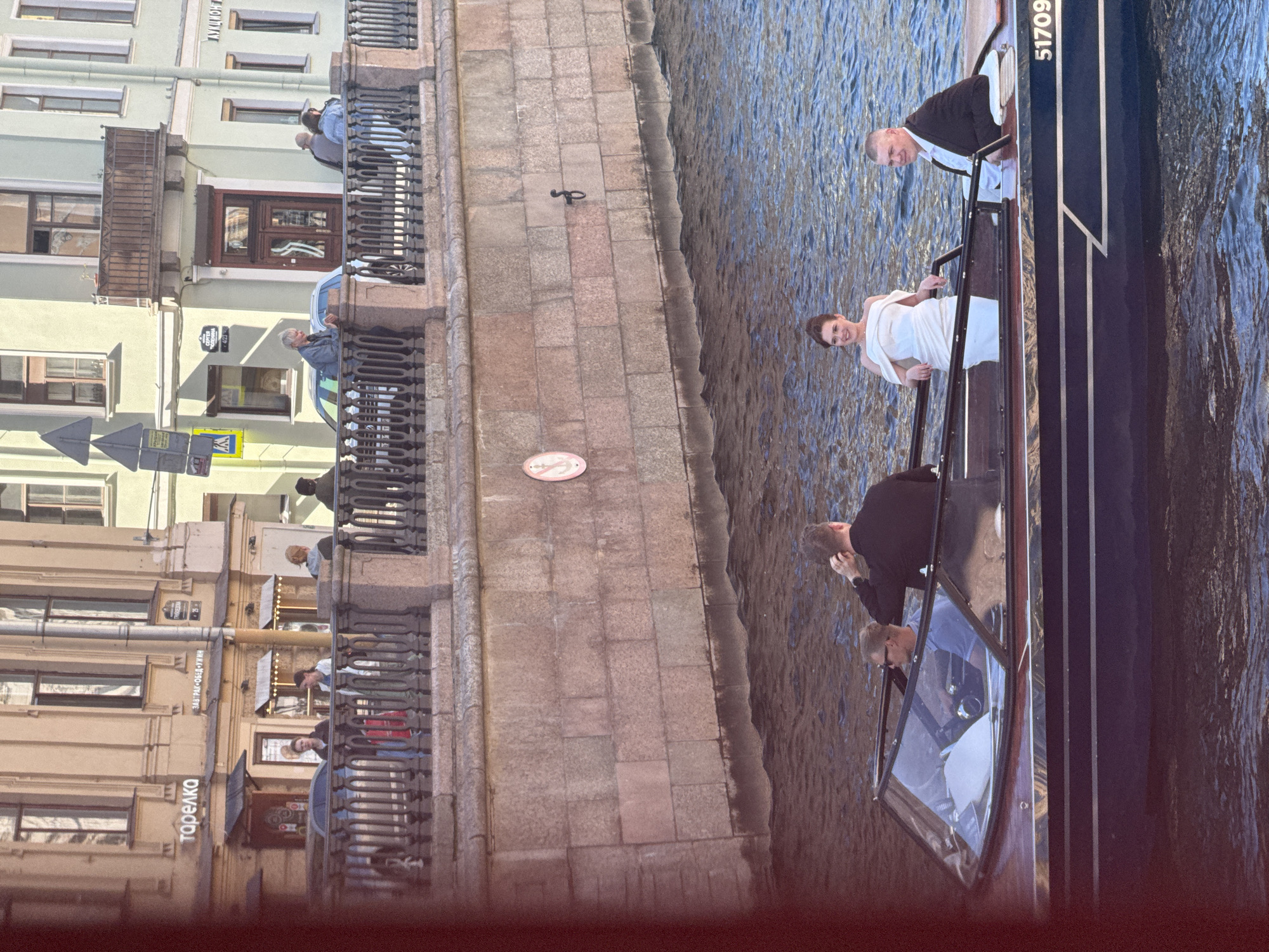 In Saint Petersburg, Russia, on a sunny day, a motorboat navigates a canal. Onboard are four individuals. A man in a black jacket and a woman in a white off-the-shoulder dress are positioned at the stern. In the middle of the boat, a man wearing a blue shirt and sunglasses is operating the vessel. Towards the bow, another man in a black jacket is visible. The boat displays the number "51709" on its side. Along the canal, buildings with cream and green facades line the embankment, featuring balconies and windows. A black metal fence runs along the top of the embankment. One building has signage that reads "тарелка" and "ЗАВТРАК-ОБЕД-УЖИН."