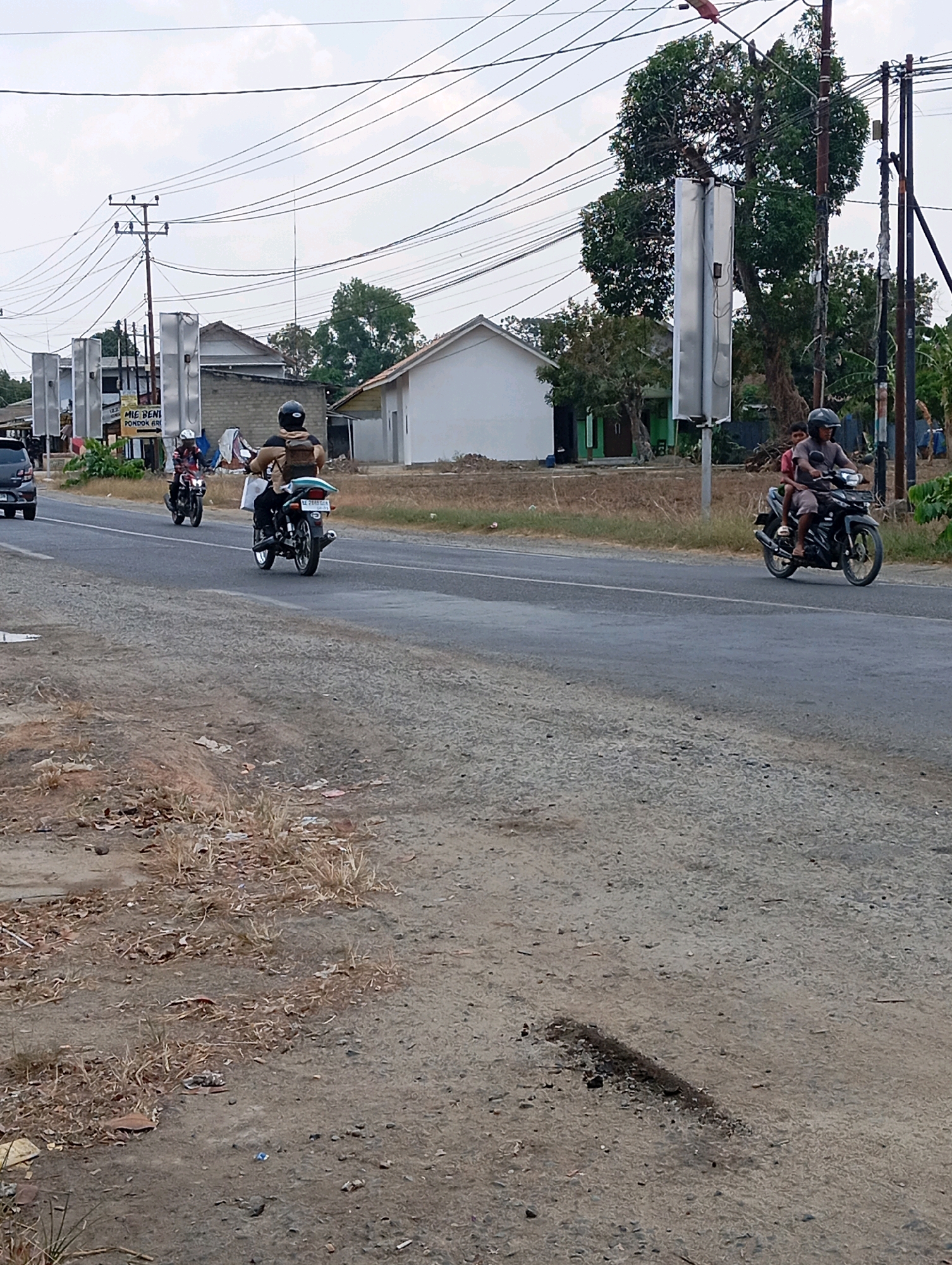 The image shows a street scene in Leman Benawi, Indonesia, under a cloudy sky. In the foreground, a dirt and gravel roadside with dry grass and scattered debris is visible. A paved road runs through the middle ground, with several motorcycles traveling on it. The first motorcycle, closer to the camera and on the left side of the road, has a rider wearing a helmet and a backpack. Another motorcycle, further down the road and on the right, carries two people, both wearing helmets. In the background, there are buildings, including a white-colored structure and a building with a brick wall. Electrical poles with numerous wires crisscross the sky above. Trees and foliage are present on both sides of the road, with one prominent large tree with broad leaves behind a tall, silver, rectangular structure. The overall impression is of a rural or suburban roadside environment.