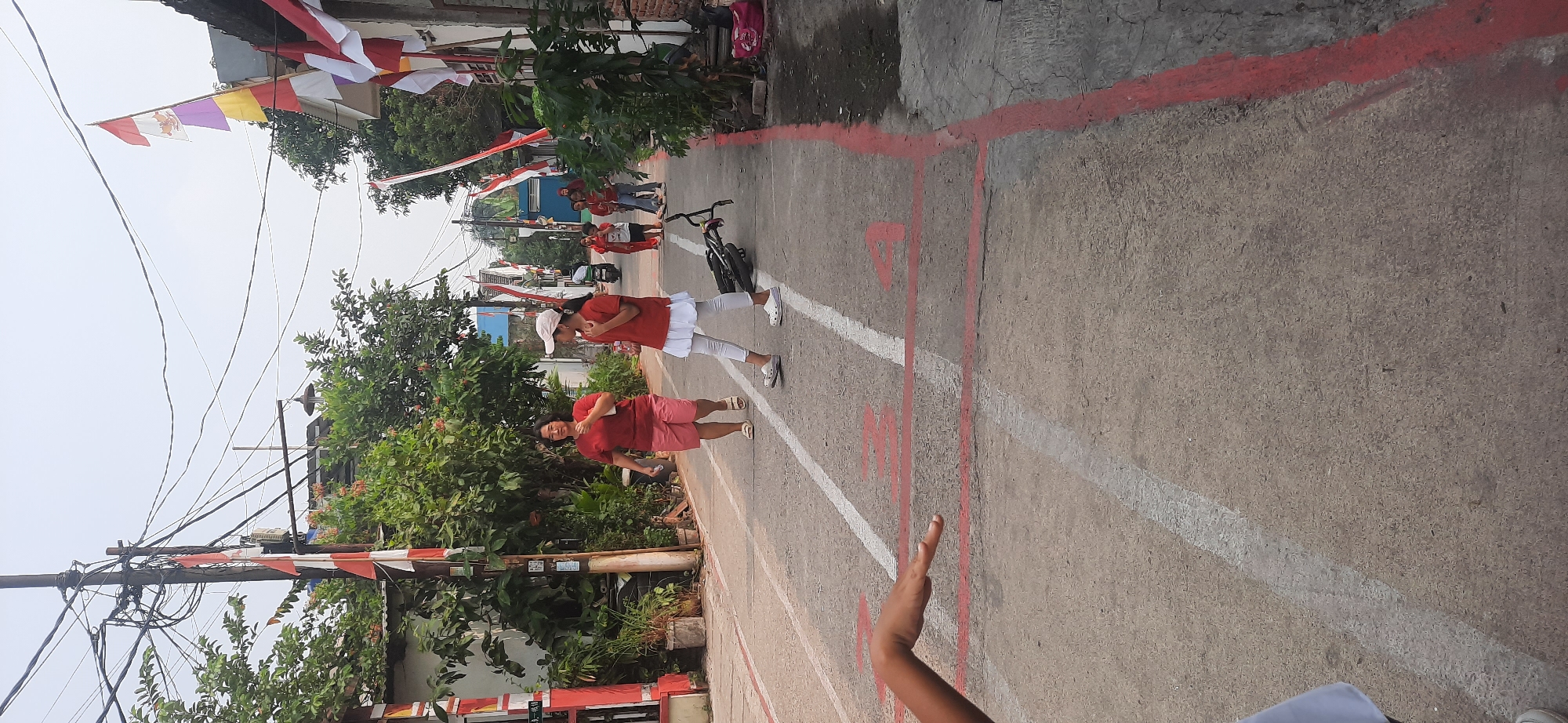In Cisauk, Indonesia, on a paved street adorned with red and white Indonesian flags, a group of people are participating in an outdoor event. Two women in red shirts and shorts walk along the street, one appearing to be the focal point of the image, while a young girl in a red shirt and white skirt stands nearby, facing away from the camera. A bicycle lies on its side in the street between them. Other individuals, including children and a person on a motorcycle, are visible in the background, suggesting a community gathering or celebration. The street is marked with white and red lines, possibly indicating lanes or a racecourse, and bordered by houses and lush green foliage. The sky is overcast, casting a diffused light on the scene.