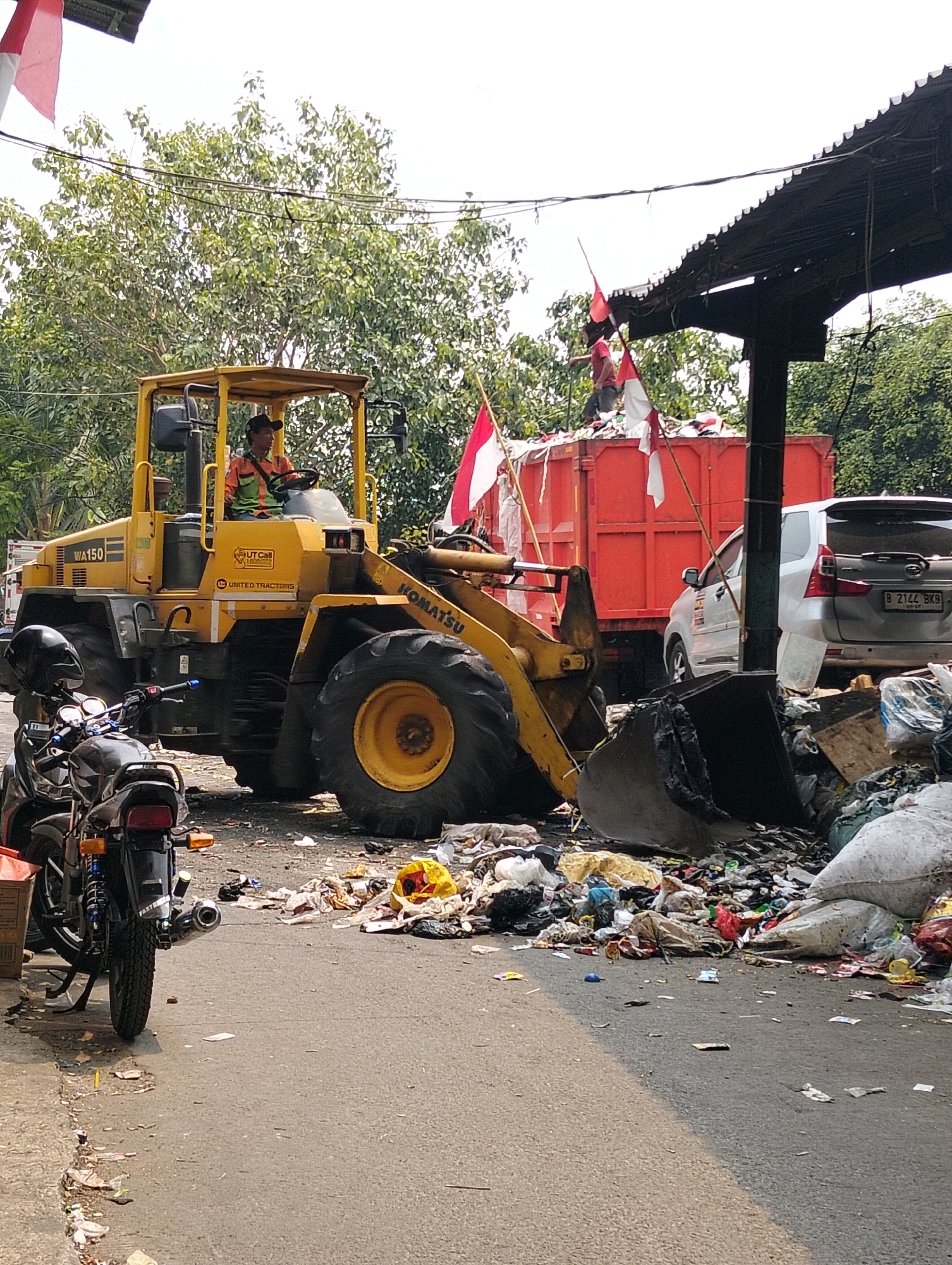 In a city street scene in West Jakarta, Indonesia, a yellow Komatsu WA150 wheel loader is positioned in front of a large red dump truck filled with trash. A man wearing an orange and green safety vest and a black cap is operating the wheel loader. The loader's bucket is lowered and appears to be collecting or moving debris from a large pile of garbage on the right side of the frame. Bags of trash and loose refuse are scattered across the asphalt road and sidewalk area. To the left of the loader, a black motorcycle with its kickstand down is parked. In the background, trees provide shade, and a white car is visible behind the dump truck. Indonesian flags are displayed on poles, with one prominently placed above the dump truck and another visible to the left. The overall environment suggests a waste disposal or cleanup operation taking place in an urban setting.