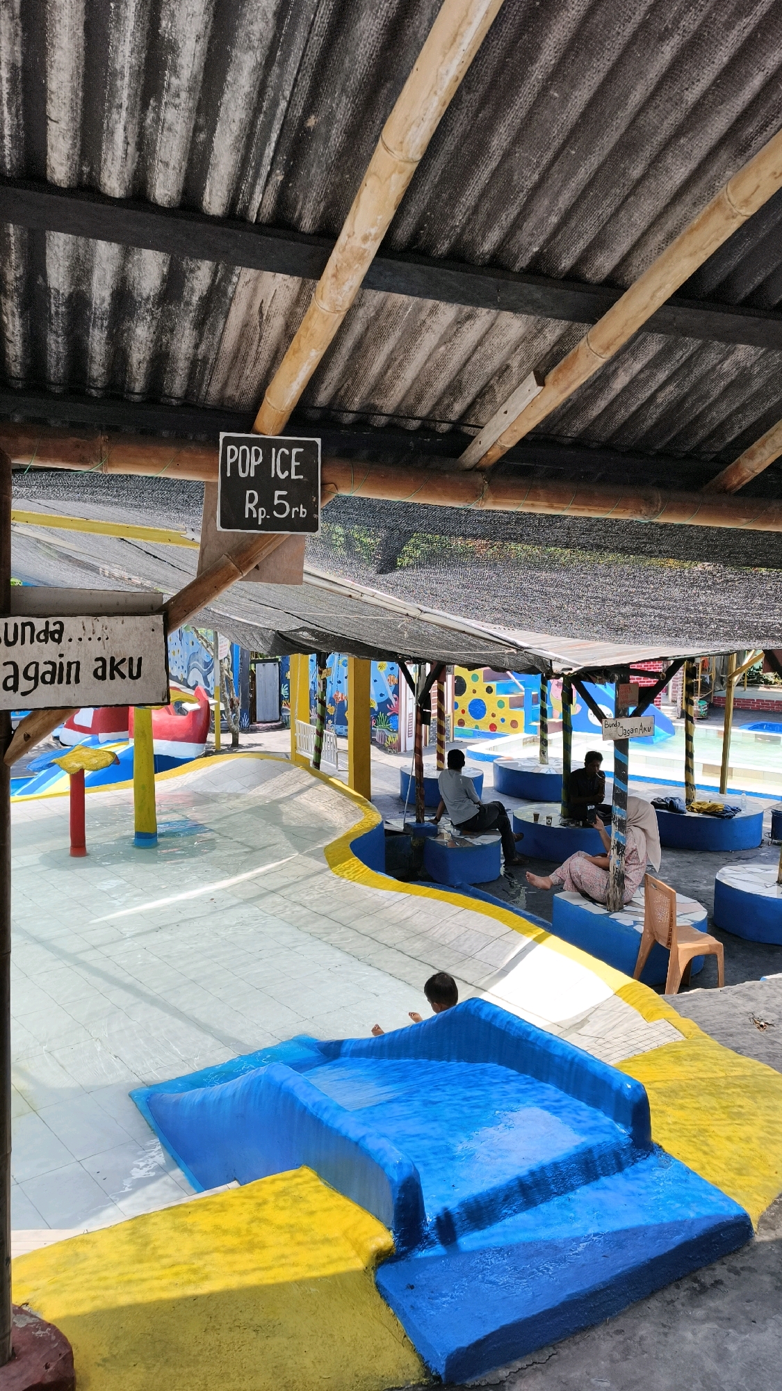 The image shows a water park area, likely in Air Paoh, Indonesia, under a textured, corrugated roof supported by bamboo poles. A sign reading "POP ICE Rp.5rb" is visible. Another sign, partially obscured, reads "Sunda...again aku".
In the foreground, a blue and yellow water slide structure is visible, with a child sitting on it. The surrounding area is a shallow pool with tiled flooring. Further back, under a black mesh covering, several people are seated on blue benches around circular tables. One person is facing away from the camera, another is looking at their phone, and a woman in a hijab is also seated.
The environment appears to be a recreational outdoor space with bright, colorful murals on the walls in the background. The overall impression is that of a casual, family-friendly water attraction.