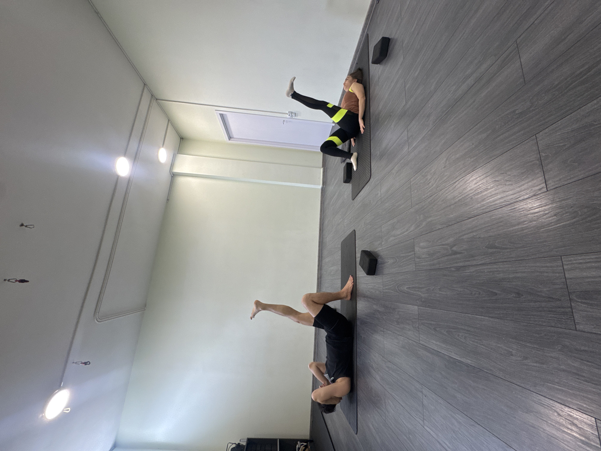 A long shot shows two people doing yoga poses on mats in a studio. The walls are light colored and the floor is dark grey. One person is wearing black shorts and a black shirt, and they are lying on their back with their legs raised. The other person is wearing black leggings with a yellow stripe, a brown shirt, and they are lying on their back with one leg raised. Each person has a block next to them on their yoga mat.