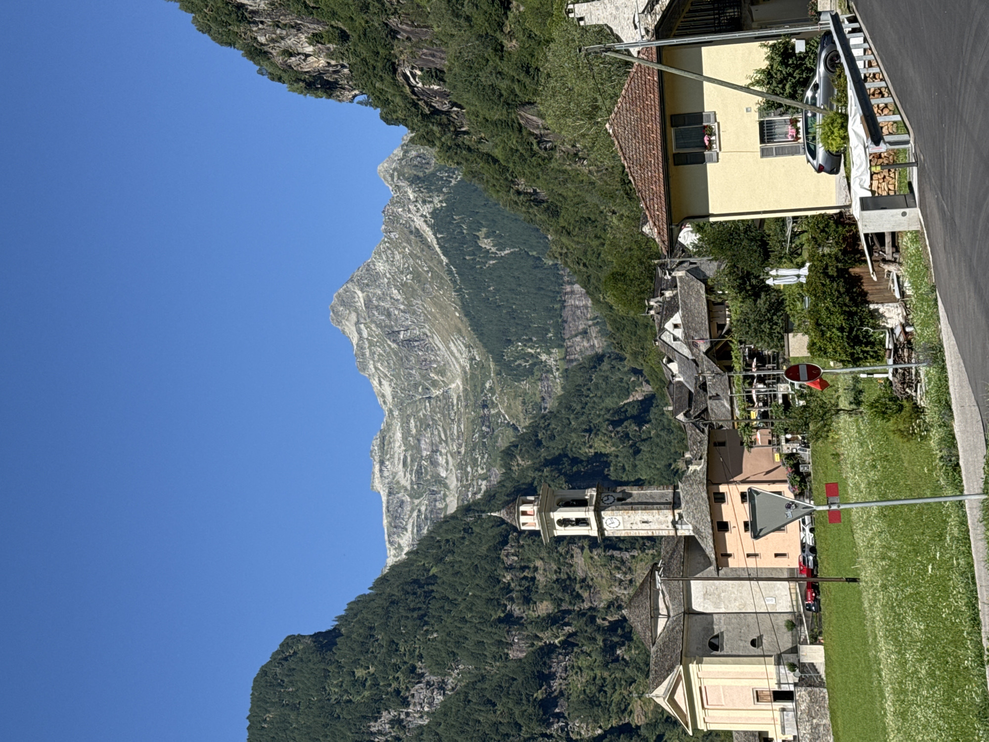 The image shows a scenic view of the town of Verzasca, Switzerland, nestled amidst lush green mountains. A prominent bell tower, part of a church, stands tall in the center of the frame, its clock face visible. Several other buildings, with their characteristic stone roofs, are scattered throughout the town. The foreground features a vibrant green meadow dotted with wildflowers, and a paved road curves through the scene. The sky above is a clear, bright blue, suggesting a sunny day. The overall impression is one of tranquility and natural beauty.
