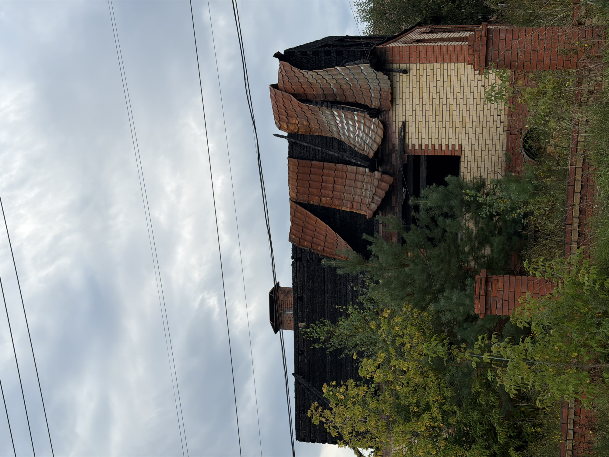 This is a full-frame, eye-level shot of a damaged house located in Chaikovsky, Russia. The house appears to have been affected by a fire, with significant damage to the roof, which is partially collapsed and has several sections of orange tiles bent and lifted. The roofing material beneath the tiles is black and appears charred. The upper walls of the house are constructed of light-colored bricks, with some sections showing signs of soot and damage. There is a brick chimney with a cap on the left side of the roof, and another brick structure, possibly a pillar or part of a fence, in the foreground on the right. Green trees and vegetation surround the house, with some foliage obscuring parts of the lower walls. The sky above is cloudy and gray. Power lines are visible in the upper portion of the image. There are no people or animals visible in the scene. The overall impression is one of disrepair and the aftermath of a fire.