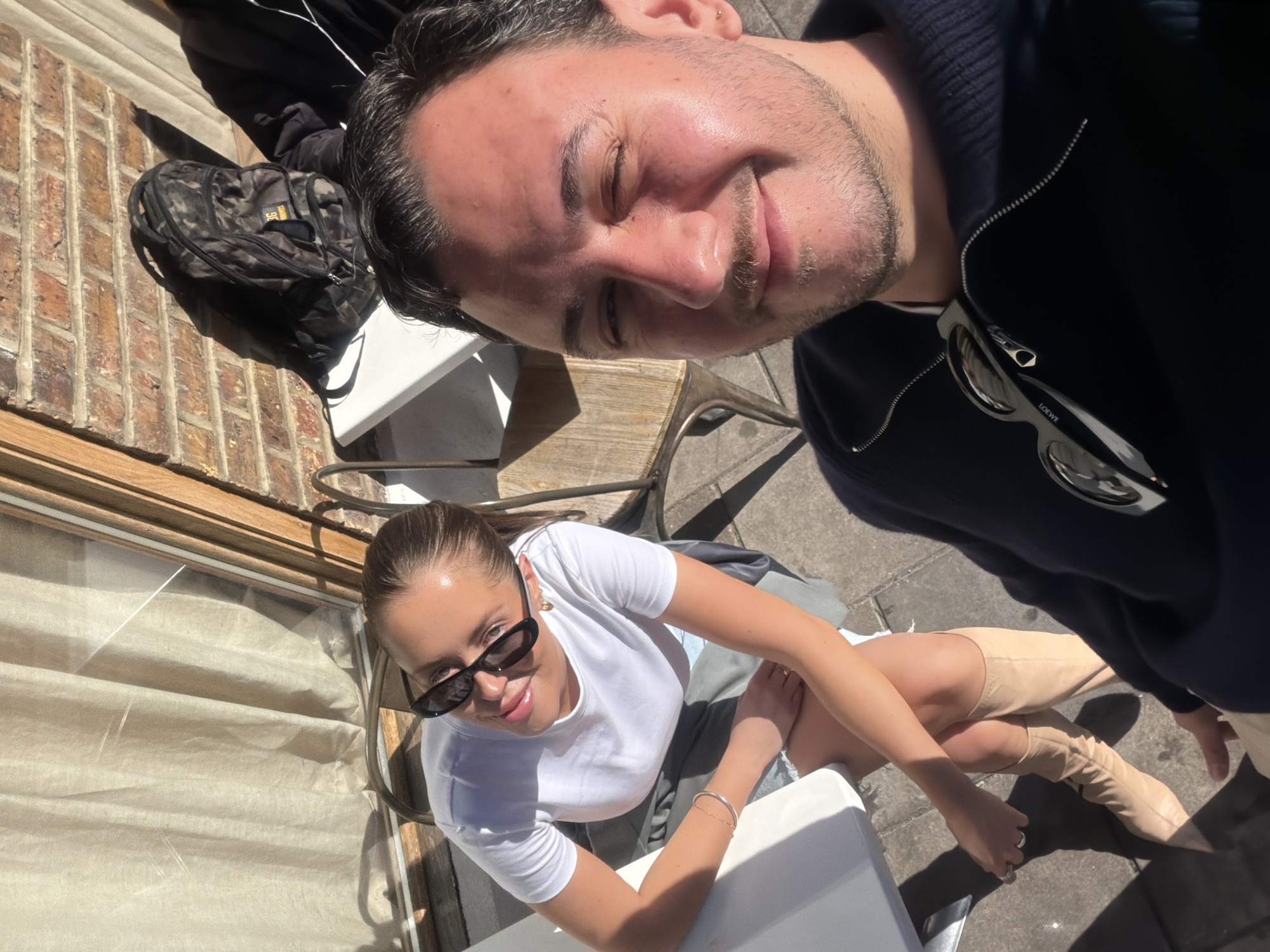 The image depicts two individuals, likely in their 20s or 30s, posing for a selfie outdoors. The man, with dark hair and a slight beard, is wearing a navy zip-up jacket with sunglasses attached, and he appears to be holding the camera. The woman, with her hair pulled back, wears sunglasses, a white t-shirt, and light-colored knee-high boots. They are seated at a table, with a backpack and chairs visible in the background. The scene is set on a brick-paved area, possibly an outdoor seating area of a café or restaurant. The light suggests it is a sunny day. The location appears to be somewhere in London based on the visible architecture, most notably the brick wall.