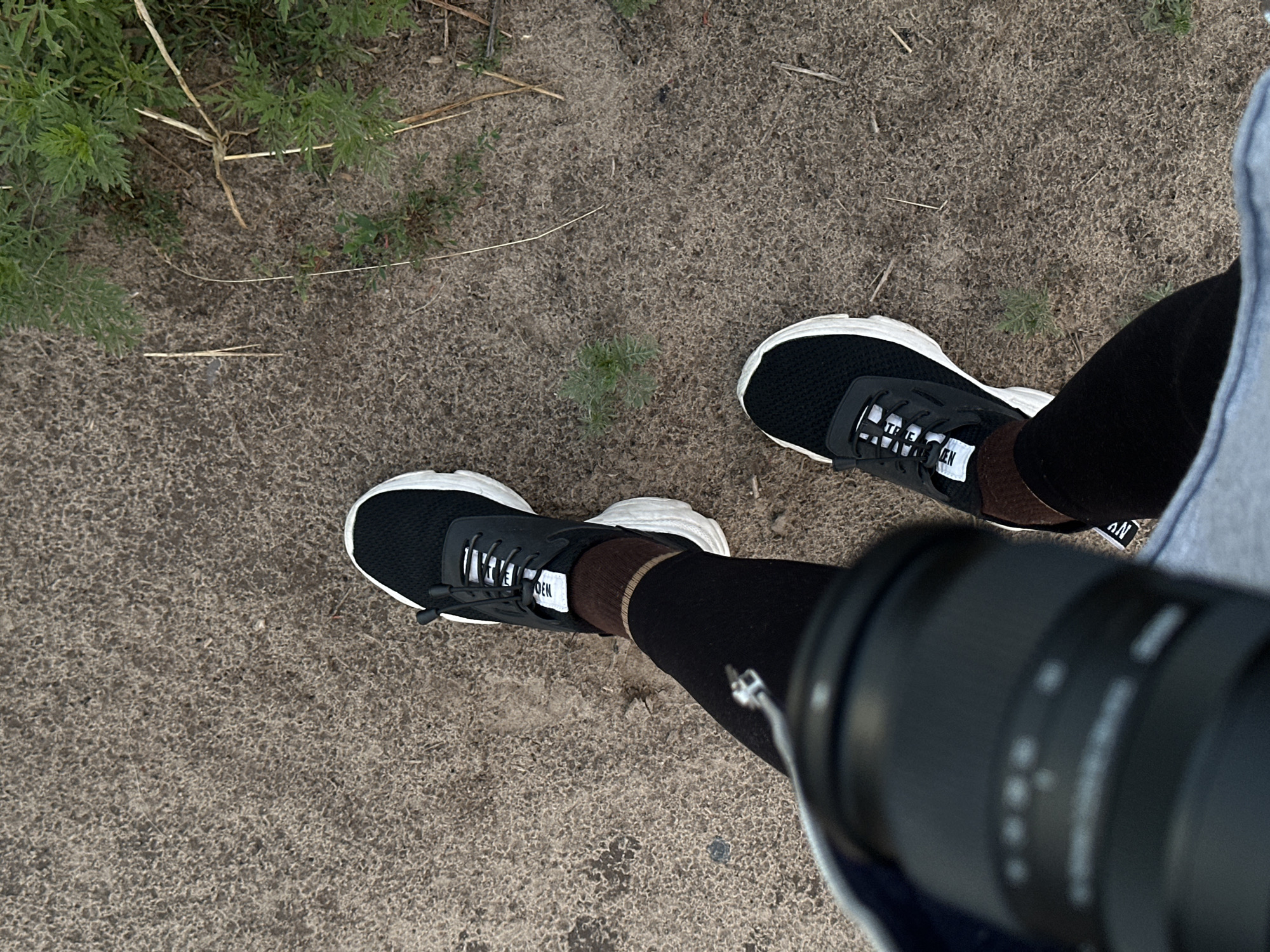 The image shows a first-person perspective of a person wearing black sneakers with white soles and black leggings. The ground is a mixture of dirt and dry grass. A camera lens is visible in the foreground, partially obscuring the lower part of the person's legs. There is also greenery from plants on the right side of the image. The scene is outdoors.