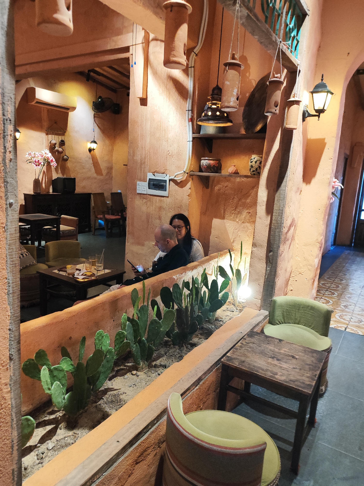 The image depicts an indoor scene that appears to be a cafe or restaurant in Da Nang, Vietnam. The decor is rustic and earthy, with prominent terracotta-colored walls and ceiling accents. In the foreground, a raised planter bed contains several green cactus plants. To the right of the planter is a small, dark wooden table with two light green chairs. One chair is partially visible, showing a patterned upholstery.
Further back in the cafe, a man and a woman are seated at a table. The man, with a shaved head and glasses, is looking down at his phone. The woman, with dark hair and glasses, is looking towards the man. Their table has drinks and what appears to be a placemat. In the background, there are more tables, chairs, and decorative elements, including hanging pottery and lights. An air conditioning unit is visible on the wall in the upper left portion of the image. The overall lighting suggests a warm and somewhat dim ambiance, typical of an evening setting in a cafe. The floor visible in the background has a tiled pattern.