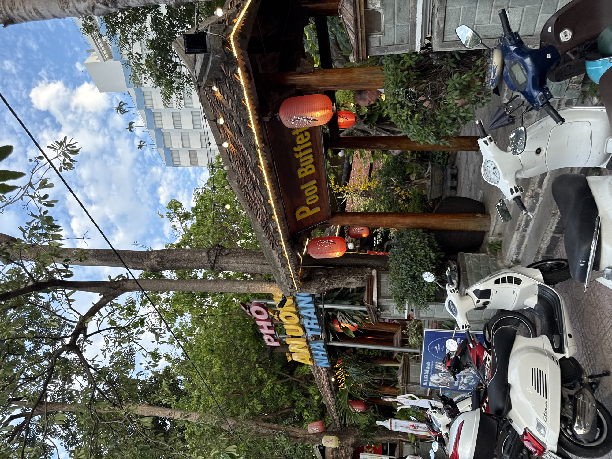 The image captures a street view in Nha Trang, Vietnam, featuring a restaurant with a prominent sign reading "PHO AN UON NHA TRANG" in vibrant colors. Adjacent to it, another sign proclaims "Pool Buffet" in yellow lettering. The establishment appears to have an outdoor dining area with a covered structure adorned with traditional tiled roofing and illuminated by string lights. Several red paper lanterns hang from the roof and trees, adding to the ambiance. A man is partially visible behind some bushes, looking to his right. In the foreground, a collection of scooters, primarily white and one dark blue, are parked. The background shows lush green trees and a modern white building with blue accents, topped with palm trees. The sky is partly cloudy.