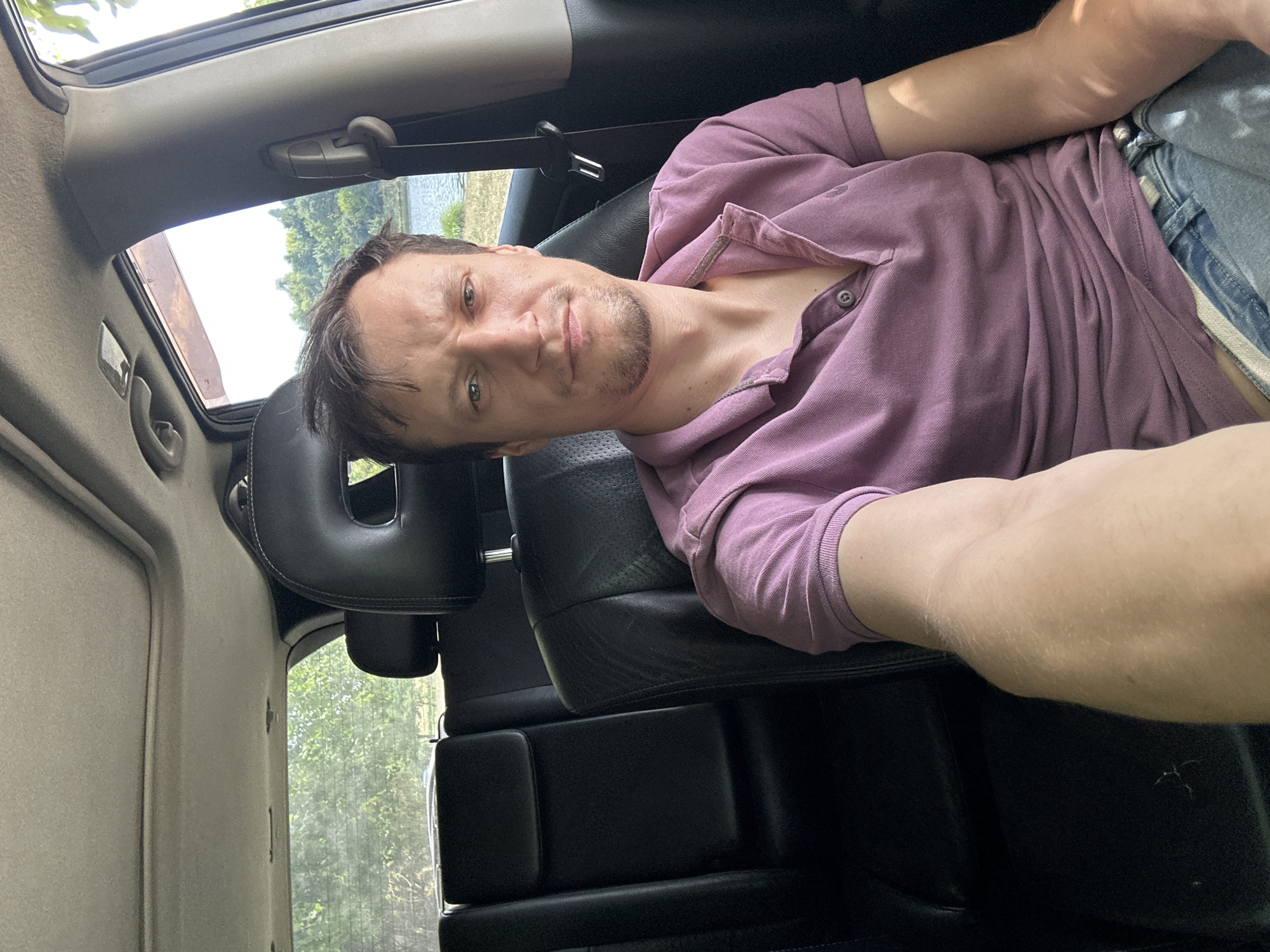 A man is sitting in the driver's seat of a car, with the camera facing him. He is wearing a pink polo shirt and blue jeans. The car's interior is visible, including the black leather seats and a sunroof. Outside the car, trees and a body of water can be seen. The image appears to be taken in Makiv, Ukraine.