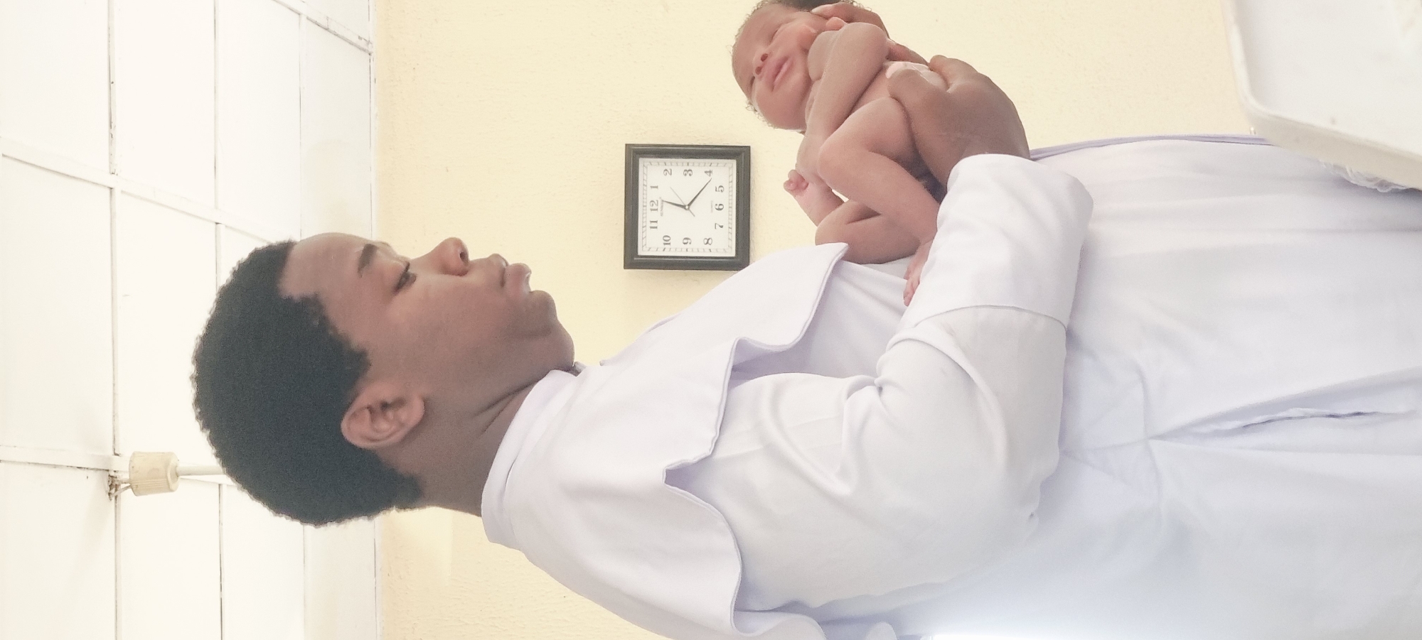 A medium shot depicts a person in a white coat holding a newborn baby against a pale yellow wall. A square-framed clock hangs on the wall, showing a time between 1:55 and 2:00. The location is unspecified in terms of it being in the city of Lagos, Nigeria. A white object is visible in the upper right corner. The person is gazing at the baby.