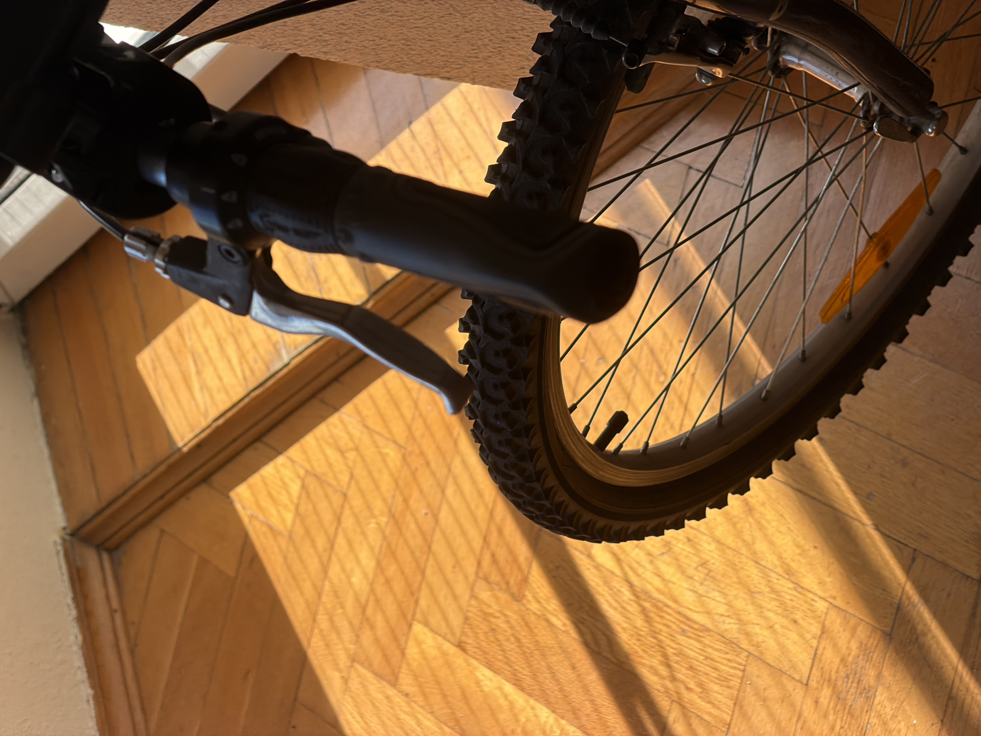 An indoor, close-up, eye-level shot focuses on the handlebars and front wheel of a bicycle. The scene is illuminated by strong, angled sunlight casting prominent shadows across a wooden parquet floor. The handlebars are black and textured, with a silver brake lever visible. The front wheel features a knobby tire, silver spokes, and a portion of the rim. The wooden floor has a warm, honey-toned finish and is laid in a herringbone pattern. The background is out of focus, but shows a white wall and a dark brown wall or object. The overall impression is one of domestic stillness, with the bicycle resting indoors.