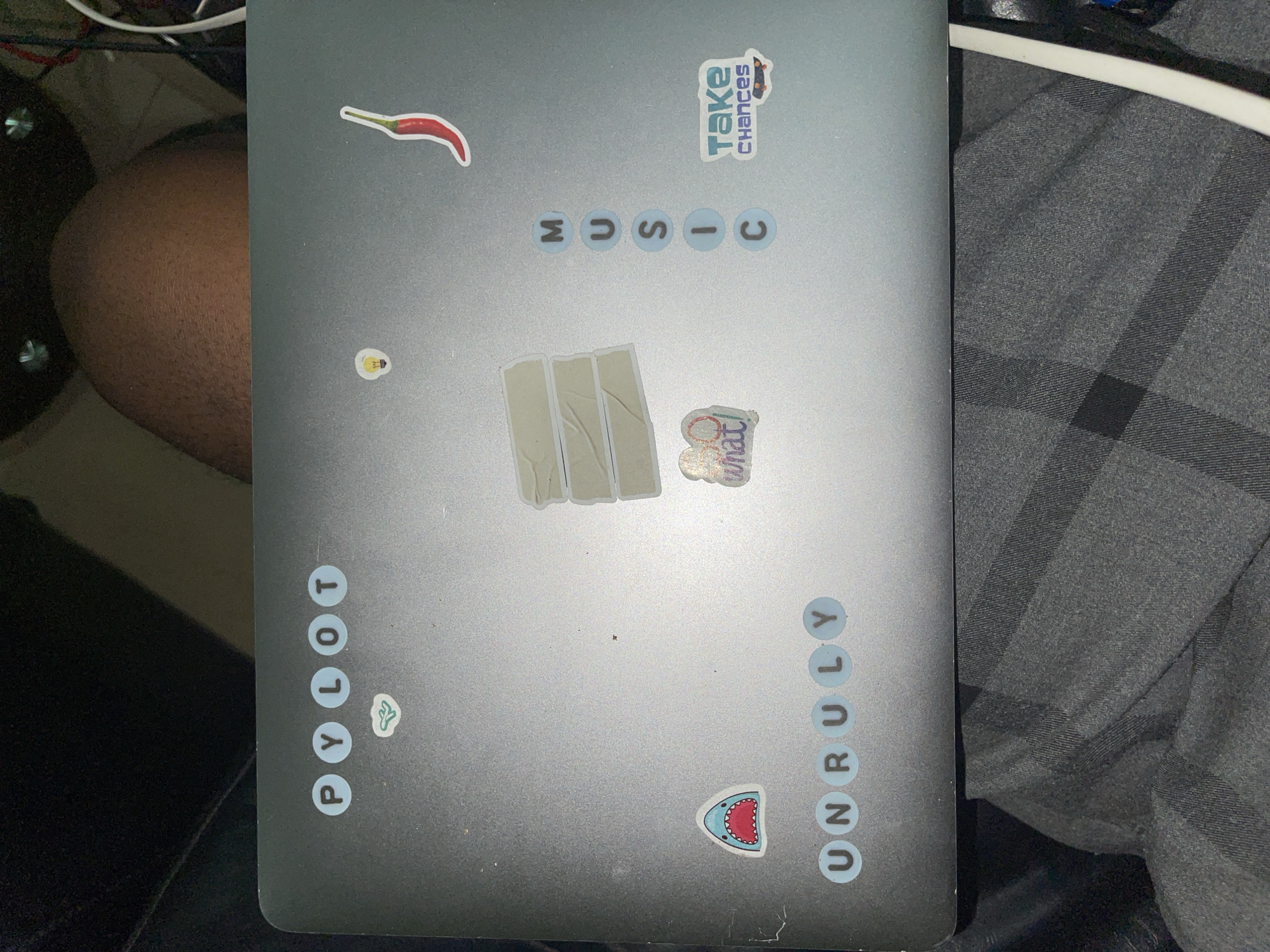 A top-down view of a closed, silver laptop covered in stickers. The stickers include a red chili pepper, the word "MUSIC" spelled with round stickers, the words "take CHANCES", three white strips, a "what!" sticker, a lightbulb, the word "PYLOT" spelled vertically with round stickers, a clover, a shark head, and the word "UNRULY" spelled vertically with round stickers. The laptop rests on what appears to be fabric. Part of a human thigh is visible to the left of the laptop. The location is unidentifiable as Abuja, Nigeria from the image alone.