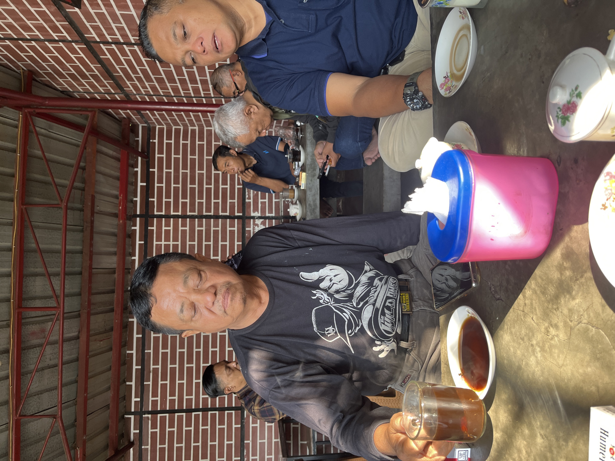 In Singosari, Indonesia, a group of men are gathered around a table at an outdoor cafe. The man in the foreground on the left is wearing a black long-sleeved shirt and grey pants, holding a glass of dark liquid, likely coffee or tea. To his right, another man in a blue polo shirt is seated, with a watch on his left wrist. Other men are visible in the background, seated at tables. The cafe features a red brick wall and a corrugated metal roof supported by red beams. The table is cluttered with cups, saucers, a tissue box, and what appears to be a smartphone. The lighting suggests it is daytime, with sunlight casting shadows.