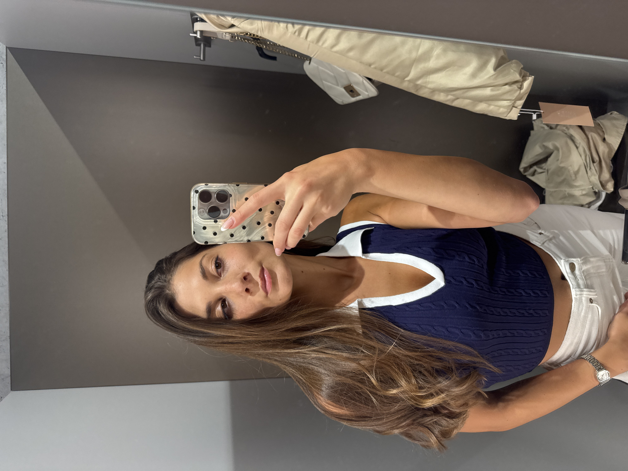 A woman is taking a mirror selfie in what appears to be a fitting room. The room has gray walls. She is wearing a navy blue top with white trim and white pants. She is holding a phone with a clear case with black dots. Her hair is long and brown. In the background, there are clothes and a white purse hanging. It is not possible to determine the specific location in Burgas, Bulgaria from this image.