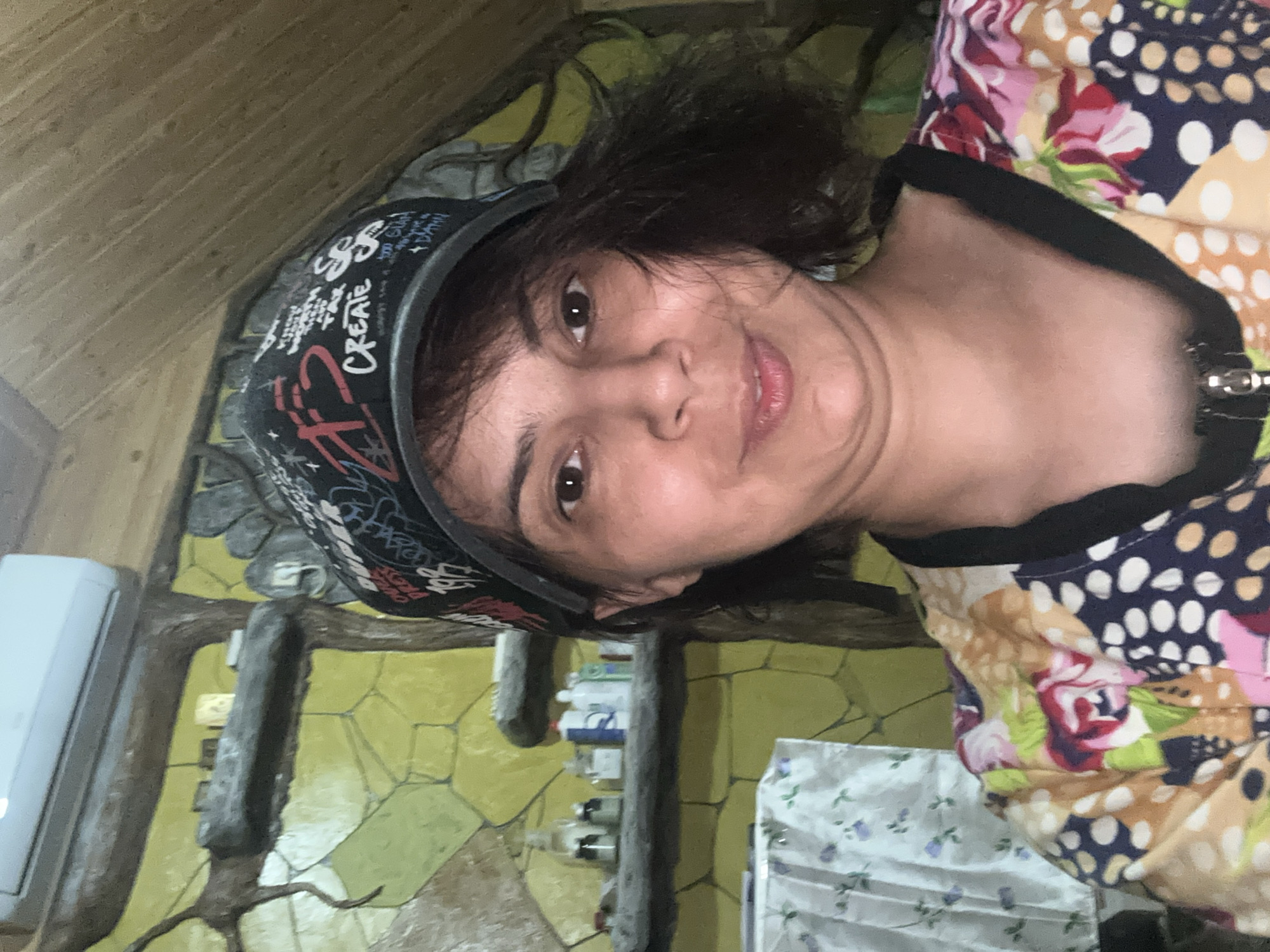 A medium shot portrait captures a woman with dark hair and a baseball cap, facing the camera. She has brown eyes and is smiling slightly. Her cap features various writings and designs in red, white, and black. She is wearing a colorful shirt with a floral and polka dot pattern. In the background, there is a shelf with bottles and jars, a textured yellow wall, and a wooden ceiling. The environment appears to be indoors, possibly a home or a workshop.