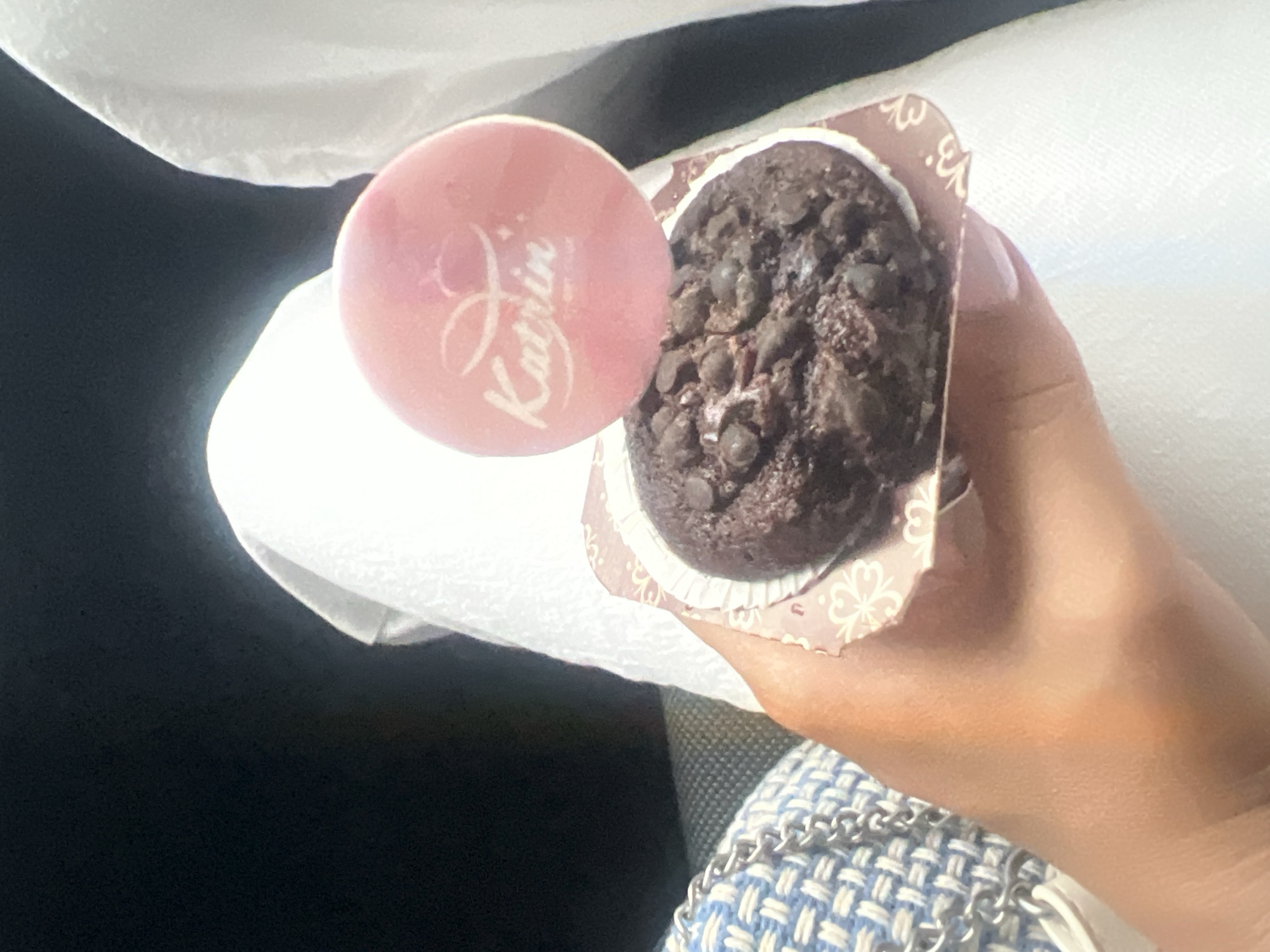 A woman's hand holds a chocolate chip muffin with a pink bonbon from the confectioner "Katrin" in the city of Plovdiv, Bulgaria. She is sitting in a car. The muffin is in a brown floral wrapper. The woman is wearing a white and blue woven jacket or purse.