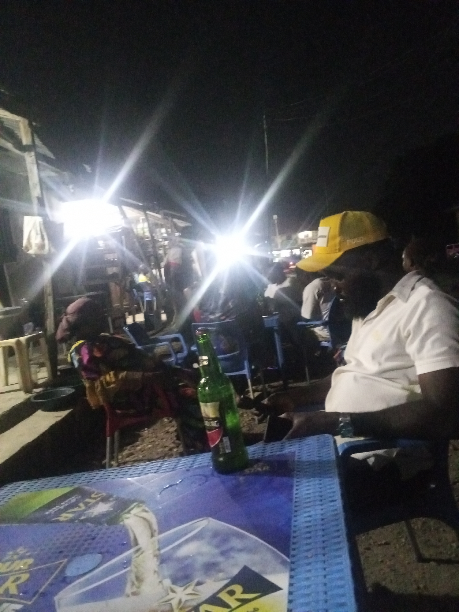 A man wearing a yellow cap and a white polo shirt sits at a blue plastic table at night in Zango-ndaji, Nigeria. A green bottle of beer, likely Star brand, stands upright on the table. In the background, there are other people sitting in chairs under bright lights, and some structures are visible. The scene appears to be an outdoor gathering or social setting at a roadside establishment.
