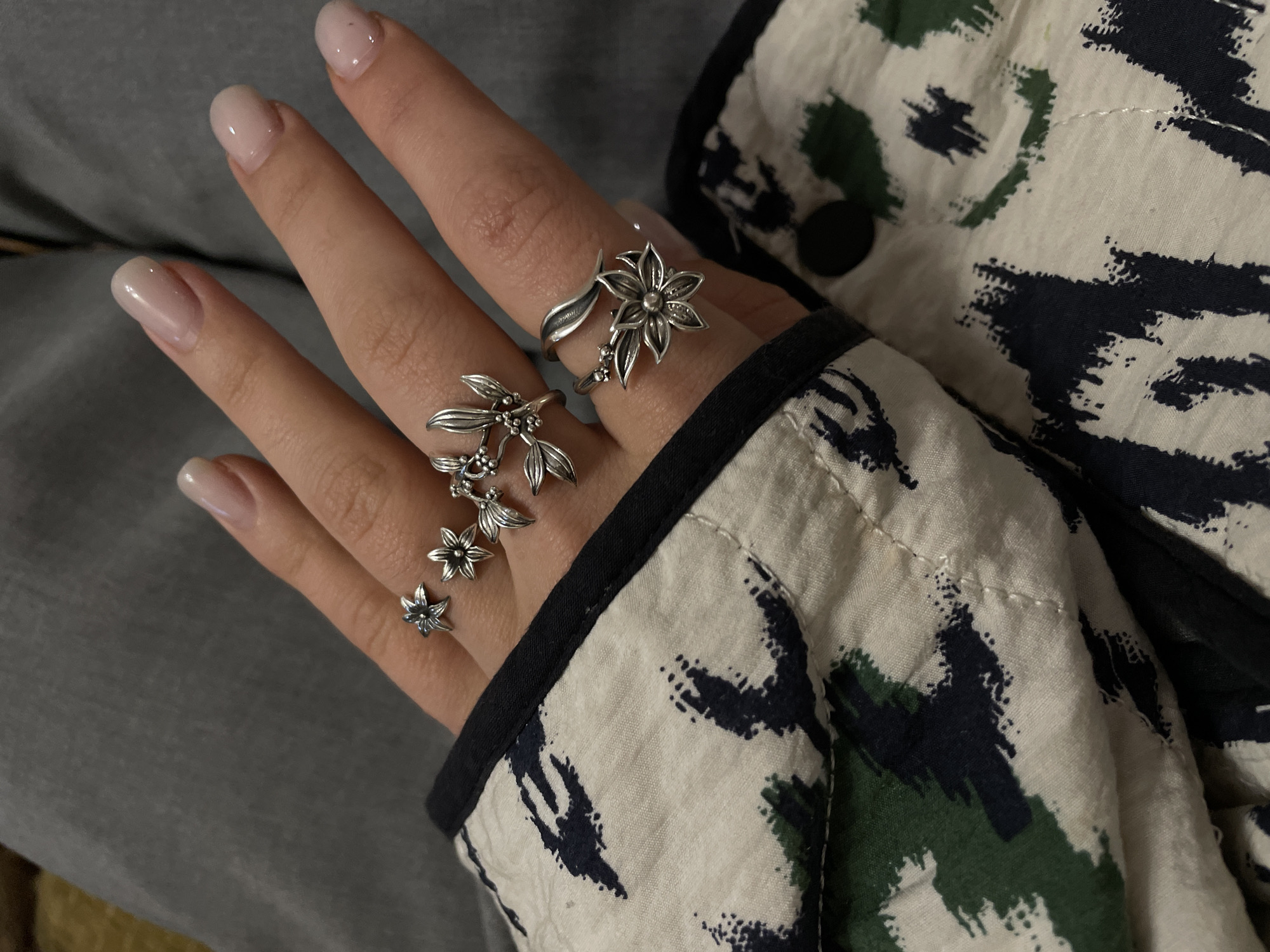 A close-up shot shows a hand adorned with two silver-colored rings. The first ring, on the index finger, features a floral design with petals extending from the center. The second ring, located on the middle finger, consists of a cascading series of three smaller flower-like elements linked together. The hand is resting against a garment with a patterned fabric that incorporates shades of cream, green, and dark blue or black, with a black trim visible along the edge. The background is a muted gray. No information suggests the image was taken in Aratashen, Armenia.