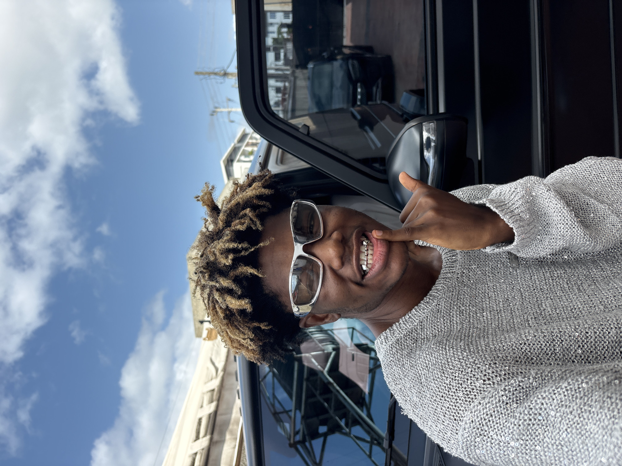 A young Black man with light brown dreadlocks, wearing silver-rimmed rectangular sunglasses and a silver, sparkly, knitted sweater, stands in front of a black Mercedes-Benz G-Wagon. He has silver grills on his teeth and is giving a hand gesture that resembles the "shaka" sign, with his thumb and pinky finger extended. His right hand is positioned near his mouth, with his index finger pointing towards his chin. The background shows a bright blue sky with scattered white clouds, a light-colored building with horizontal blinds, and power lines. Another black SUV is visible in the reflection of the G-Wagon's window. The scene appears to be an outdoor urban setting, possibly in Igboefon, Nigeria.