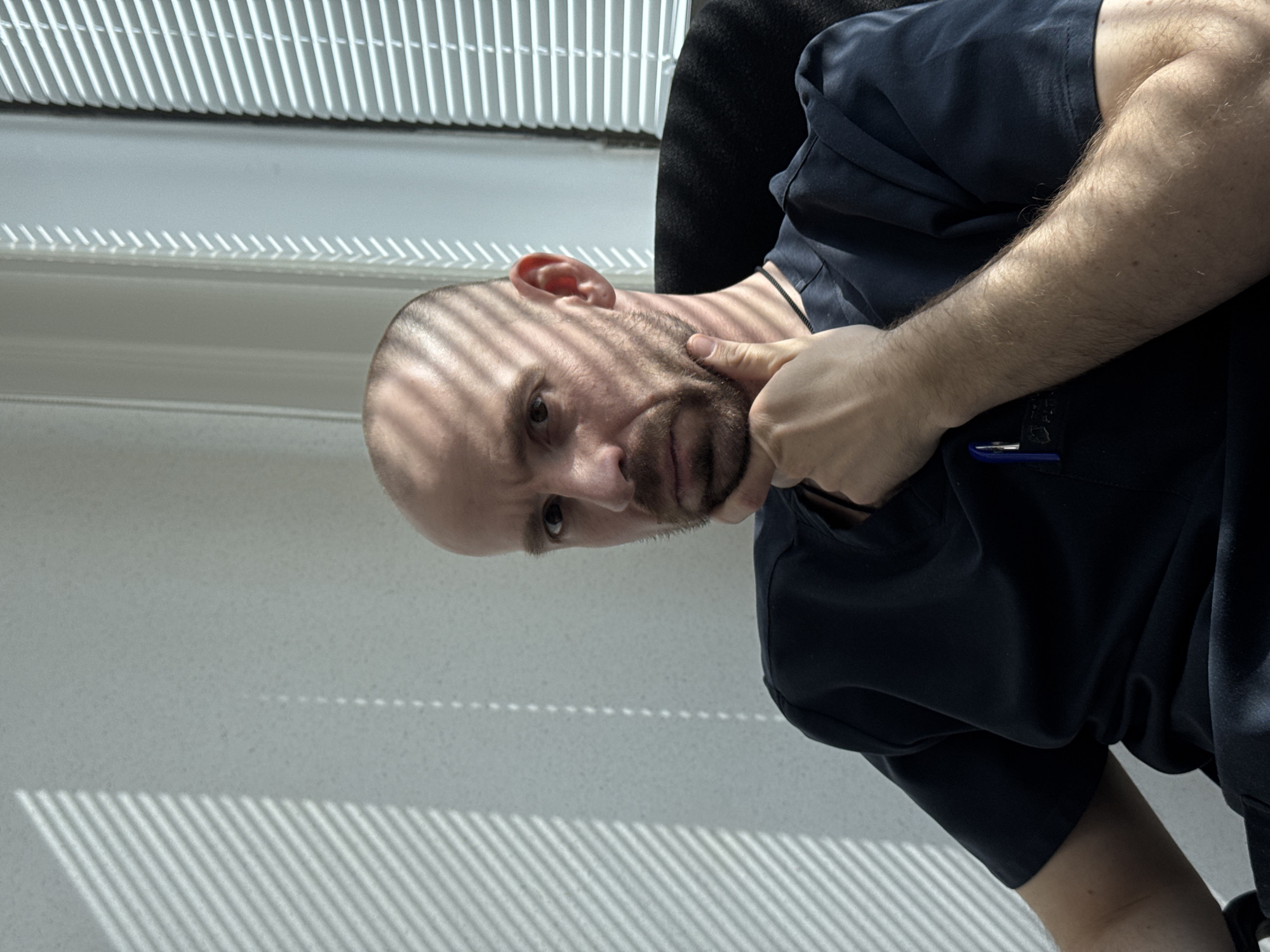 A fair-skinned man with a shaved head and a beard is sitting in a black chair near a window. He wears a dark navy shirt. His right hand is touching his beard and neck area. Light from the window blinds casts shadows across his face and the nearby wall. The location is unknown.