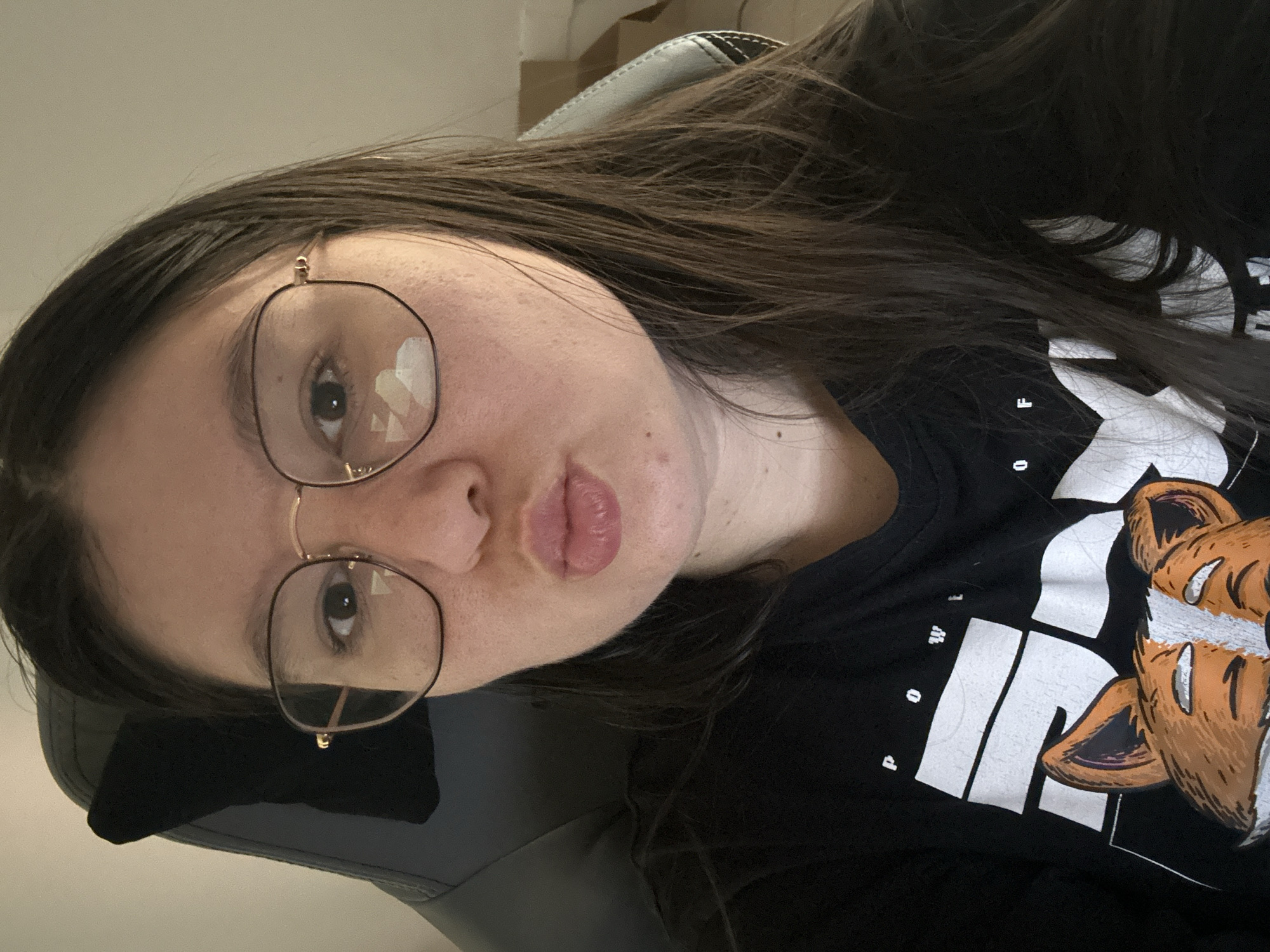 A woman with long dark hair and glasses is in the photo. She is wearing a black t-shirt with a white graphic. She has light skin and is making a kissing face. She is sitting in a chair. There is no indication of the location being Câmara de Lobos, Portugal.
