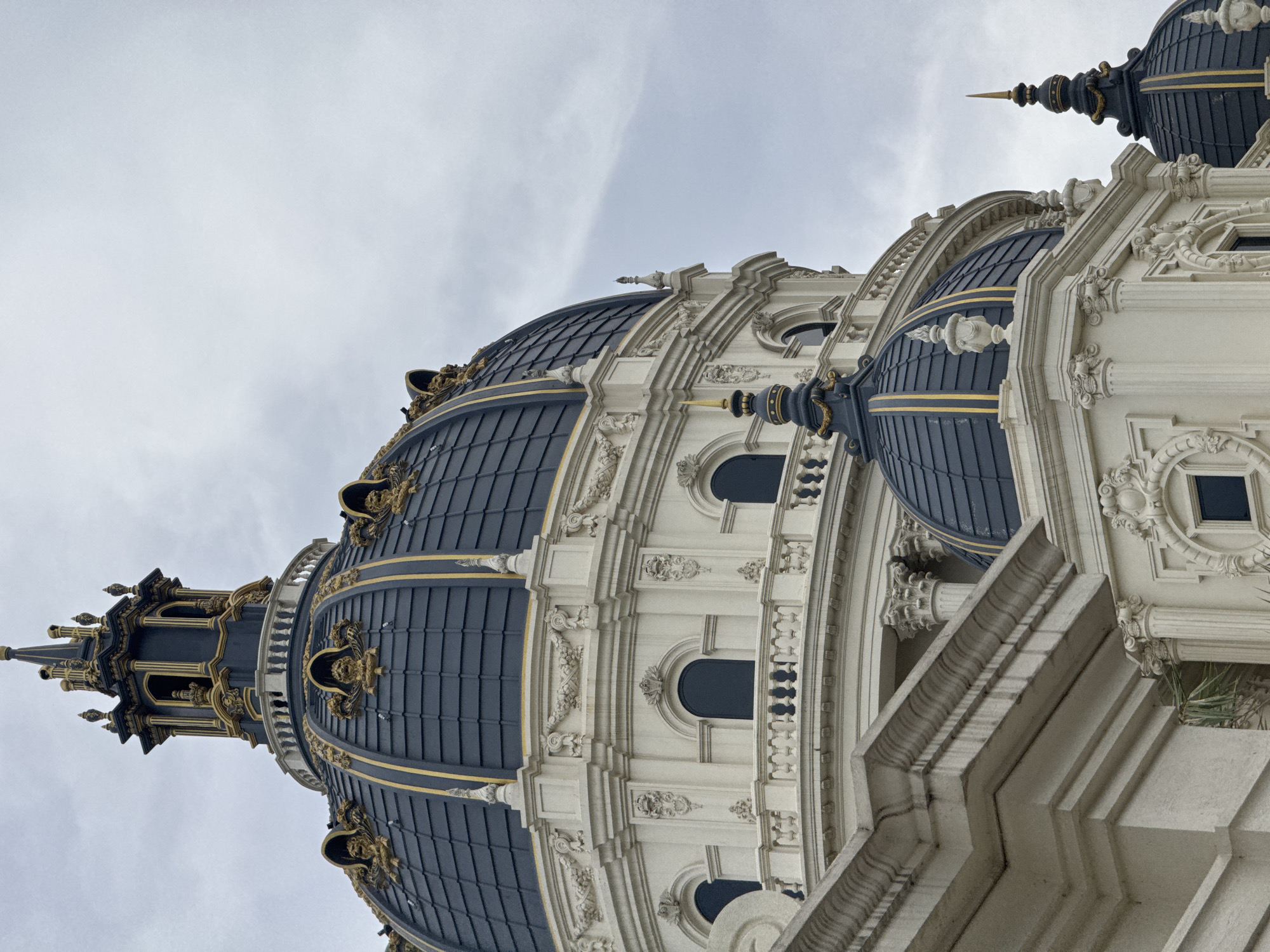 The image shows a detailed view of the opulent domes and architectural elements of a grand building in Hoa Lư Ward, Vietnam. The primary focus is on the intricate details of the domes, which are covered in dark blue tiles with ornate gold trim and decorative accents, including what appear to be lion heads or masks. A prominent central dome is topped by a multi-tiered, dark blue and gold spire. Below this, a smaller dome with a similar design is visible, also capped with a spire. The building itself is constructed of white stone with elaborate decorative carvings and columns. Several arched windows are visible, some with dark glass. A white balustrade runs along a balcony-like structure. The sky is overcast with a muted, cloudy appearance. The foreground features a stone ledge with layered molding. The overall impression is of classical, highly ornamented architecture. There are no people visible in the image.