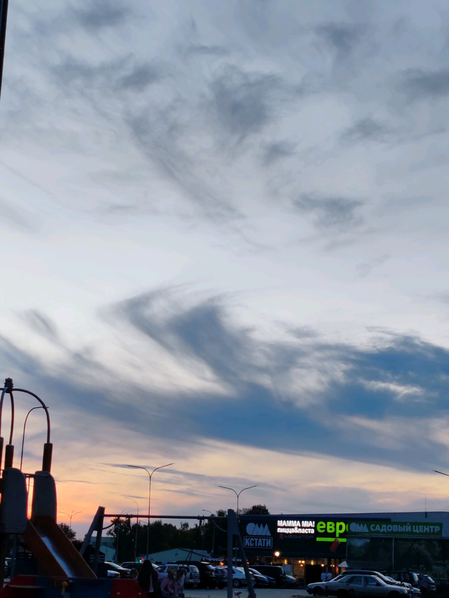 A low-angle, eye-level, vertical shot captures a playground structure in the foreground, partially obscuring a sunset sky with wispy, dramatic clouds. To the right, a strip mall is visible with a large illuminated sign that reads "MAMMA MIA! пицца&паста" and "евр" followed by "САДОВЫЙ ЦЕНТР" (Garden Center). Several parked cars are visible in front of the strip mall. A few people are standing near the cars and at the entrance to the establishment. The sky is a blend of muted grays and blues at the top, transitioning to hues of orange and yellow near the horizon, indicating dusk. Streetlights are intermittently visible against the twilight sky. The overall scene suggests an urban or suburban setting at the end of the day.