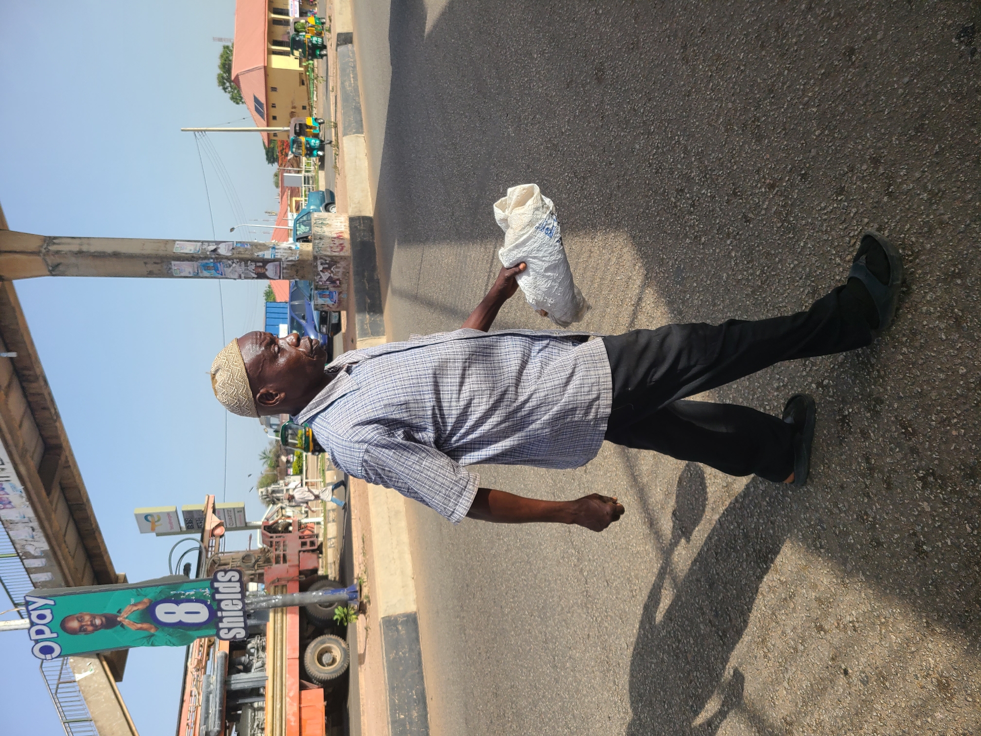 An elderly man is walking on a paved road in Gura Top, Nigeria. He is wearing a patterned kufi, a checkered short-sleeved shirt, black trousers, and dark-colored slip-on shoes. He is holding a white sack in his right hand. The road has a painted black and white curb. In the background, there is a sign advertising "OPay 8 Shields" and what looks like a petrol station price sign and a gas station or construction vehicle. Buildings and cars are visible farther in the background. The light suggests a sunny day.