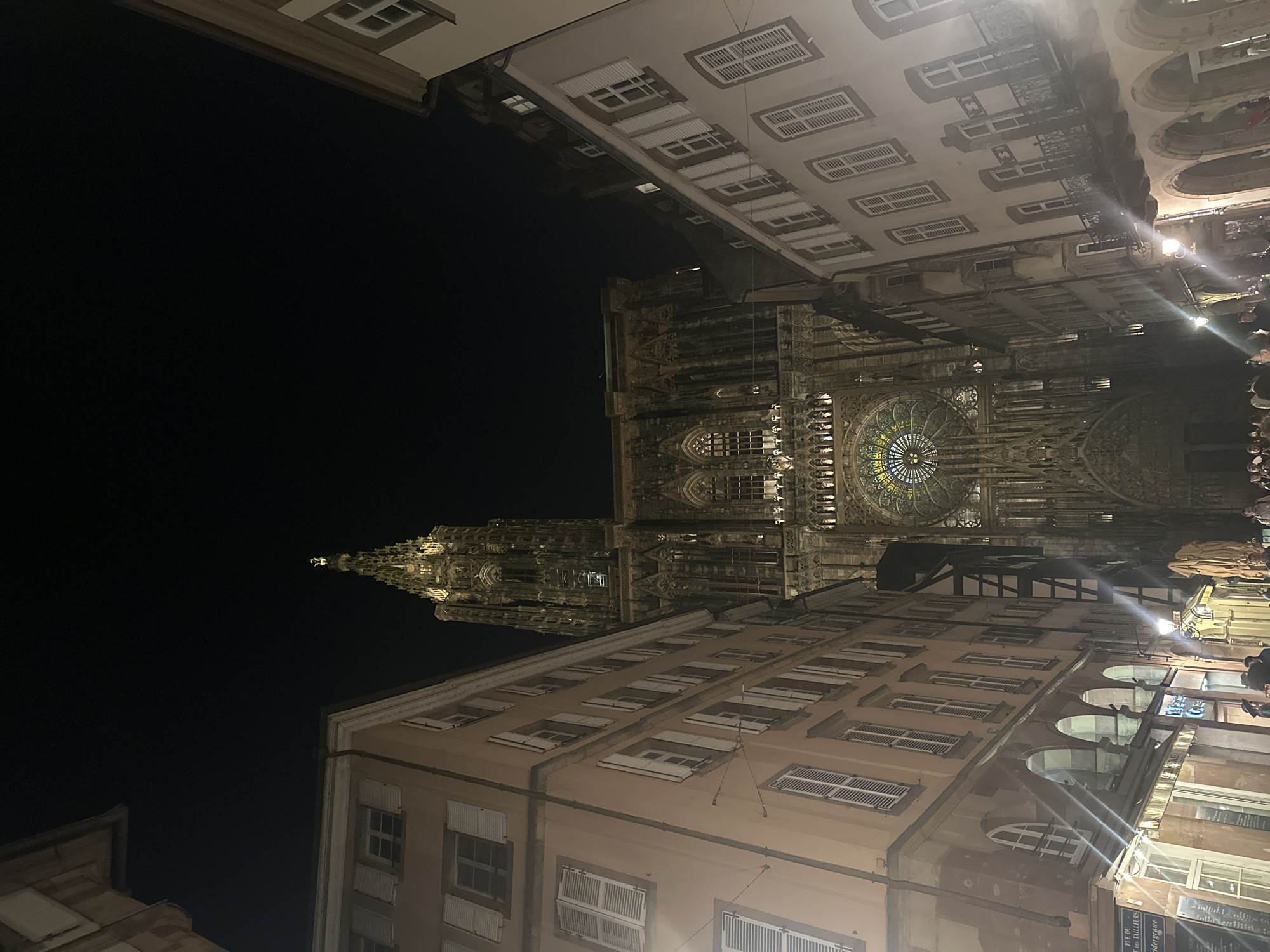 The image shows a nighttime view of the Strasbourg Cathedral, also known as Cathédrale Notre-Dame de Strasbourg, in Strasbourg, France. The cathedral's illuminated spire rises in the background, dominating the scene. The facades of surrounding buildings, with their distinctive architectural features and window shutters, frame the cathedral. Streetlights cast bright beams of light across the area. People are visible on the street level, indicating activity in the city. The overall scene captures a vibrant urban setting, with the cathedral as a focal point of cultural and historical significance.